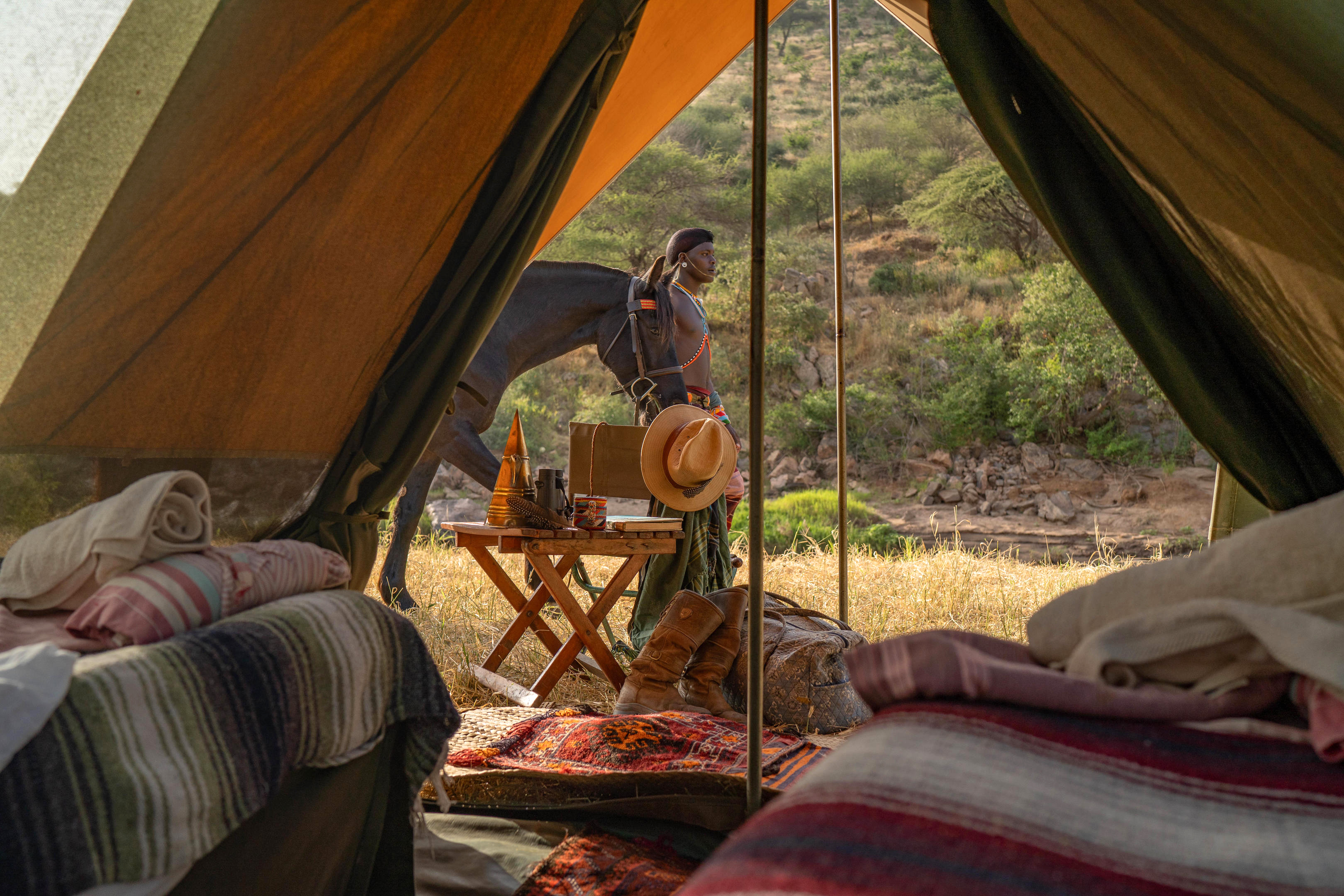 View from inside a canvas tent toward rocky hills, with a person outside near a small table and morning light.