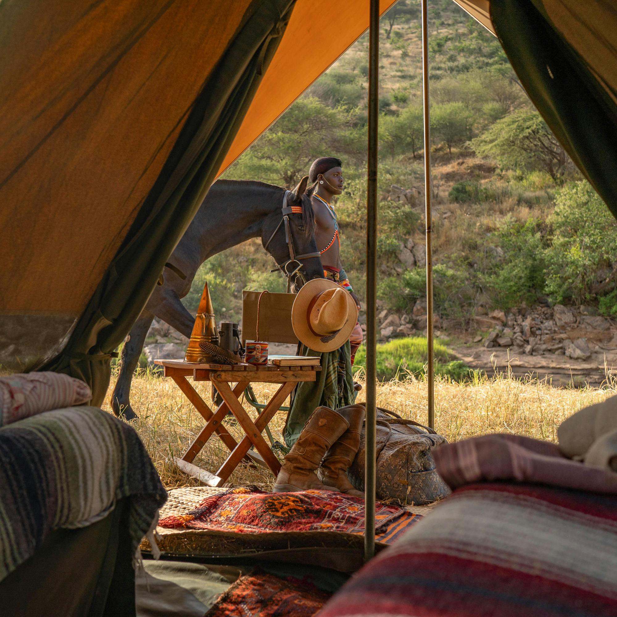 View from inside a canvas tent toward rocky hills, with a person outside near a small table and morning light.