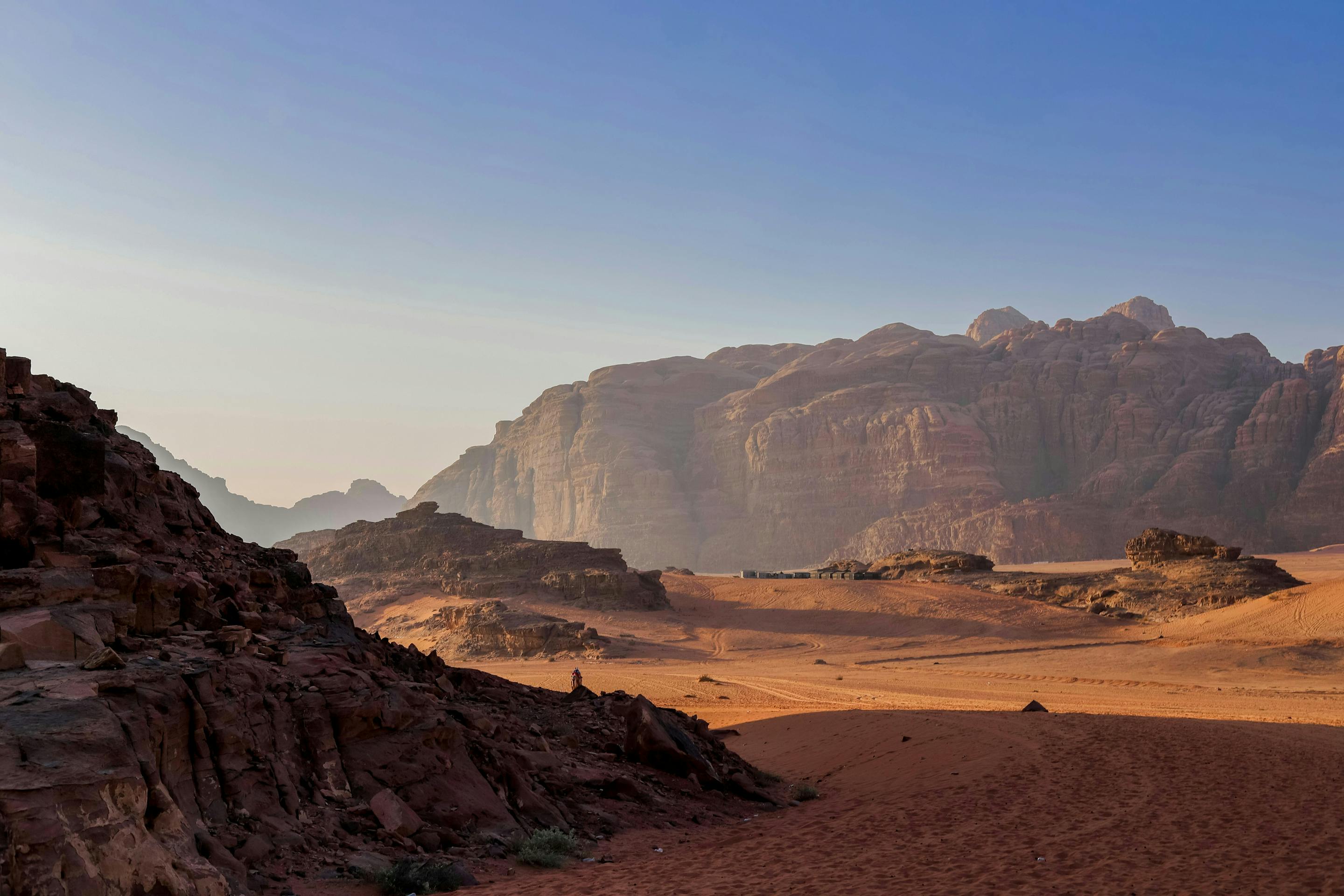 Vast desert valley with red sand and rugged cliffs, fading into haze beneath a clear sky and early light.