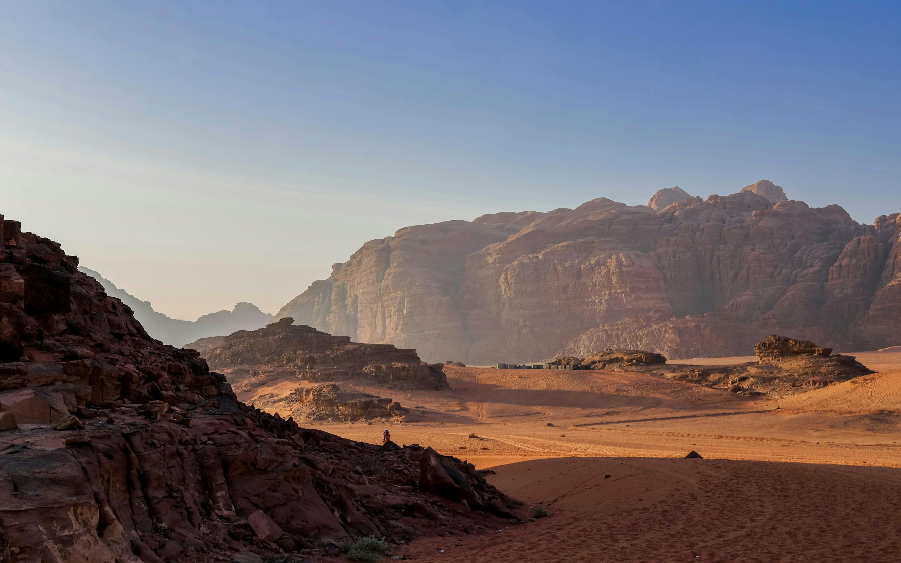 Vast desert valley with red sand and rugged cliffs, fading into haze beneath a clear sky and early light.