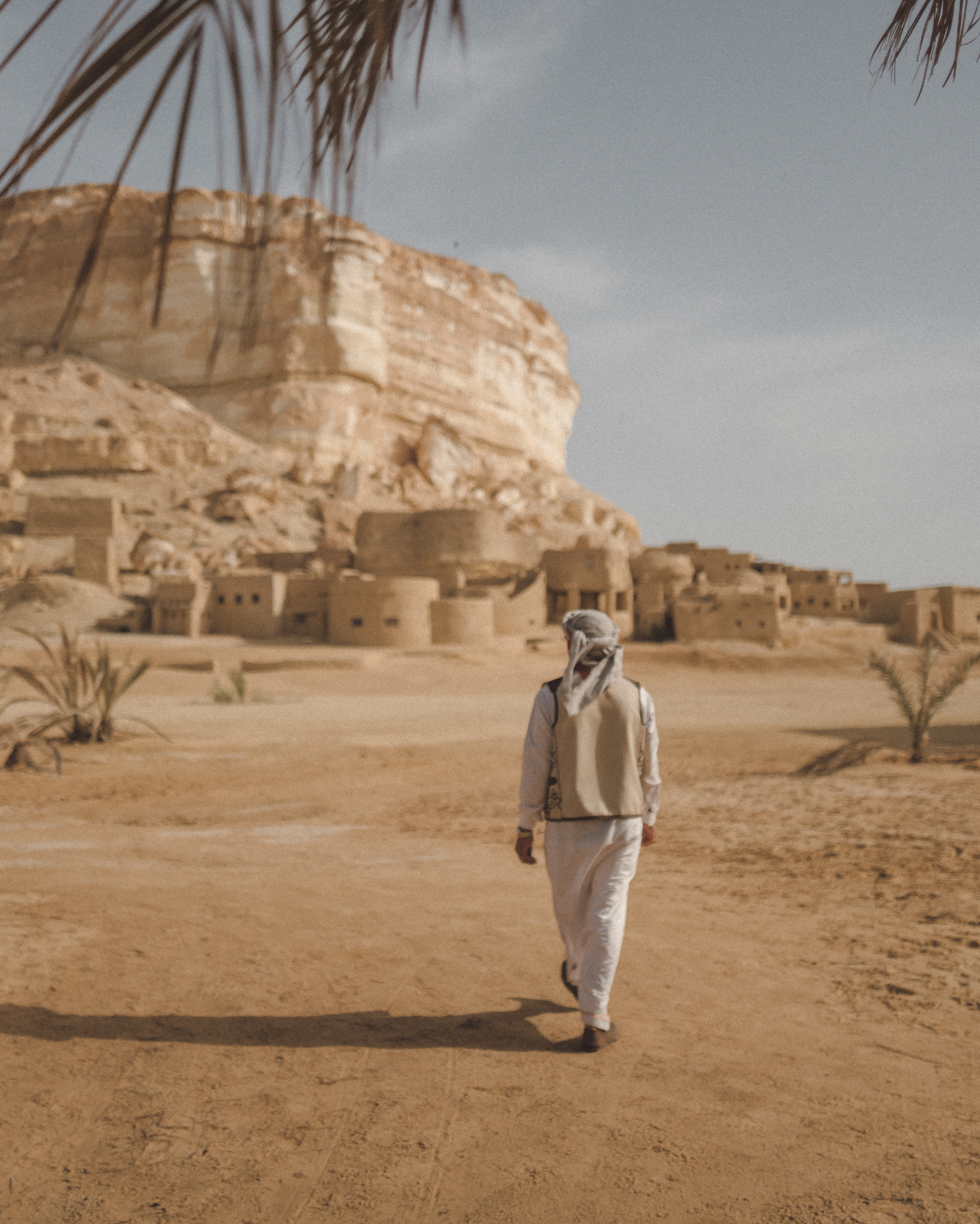 A person walks on sand near sunlit mudbrick buildings with a rocky cliff in the background.
