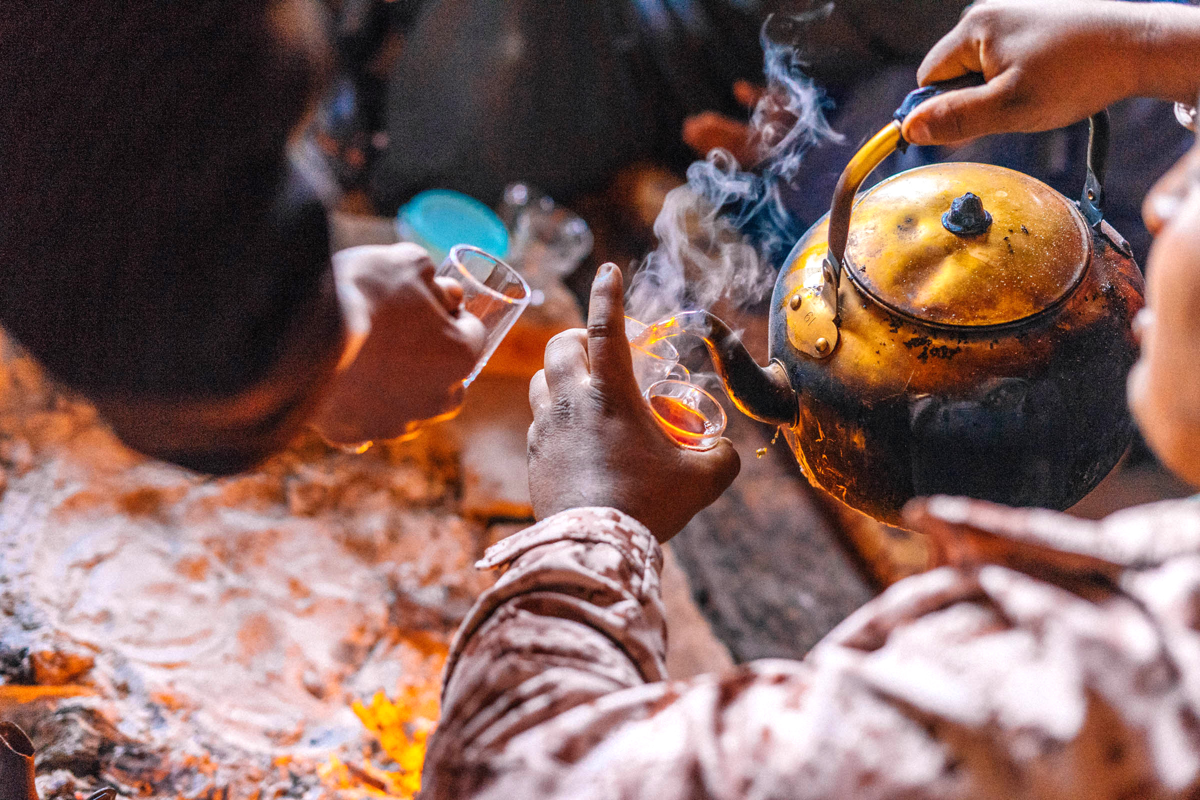 Hands pour tea from a kettle into small glasses over glowing coals.