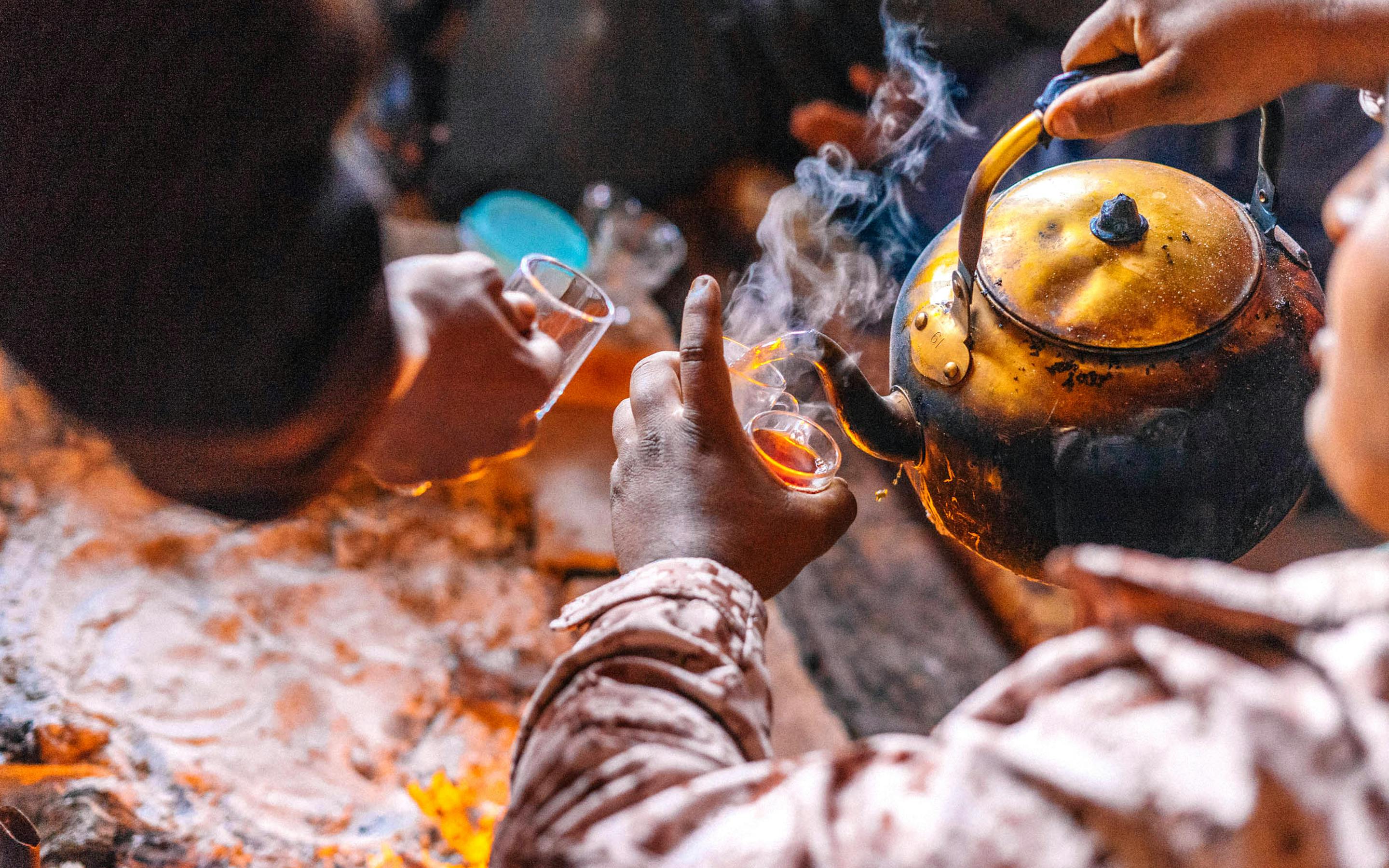 Hands pour tea from a kettle into small glasses over glowing coals.