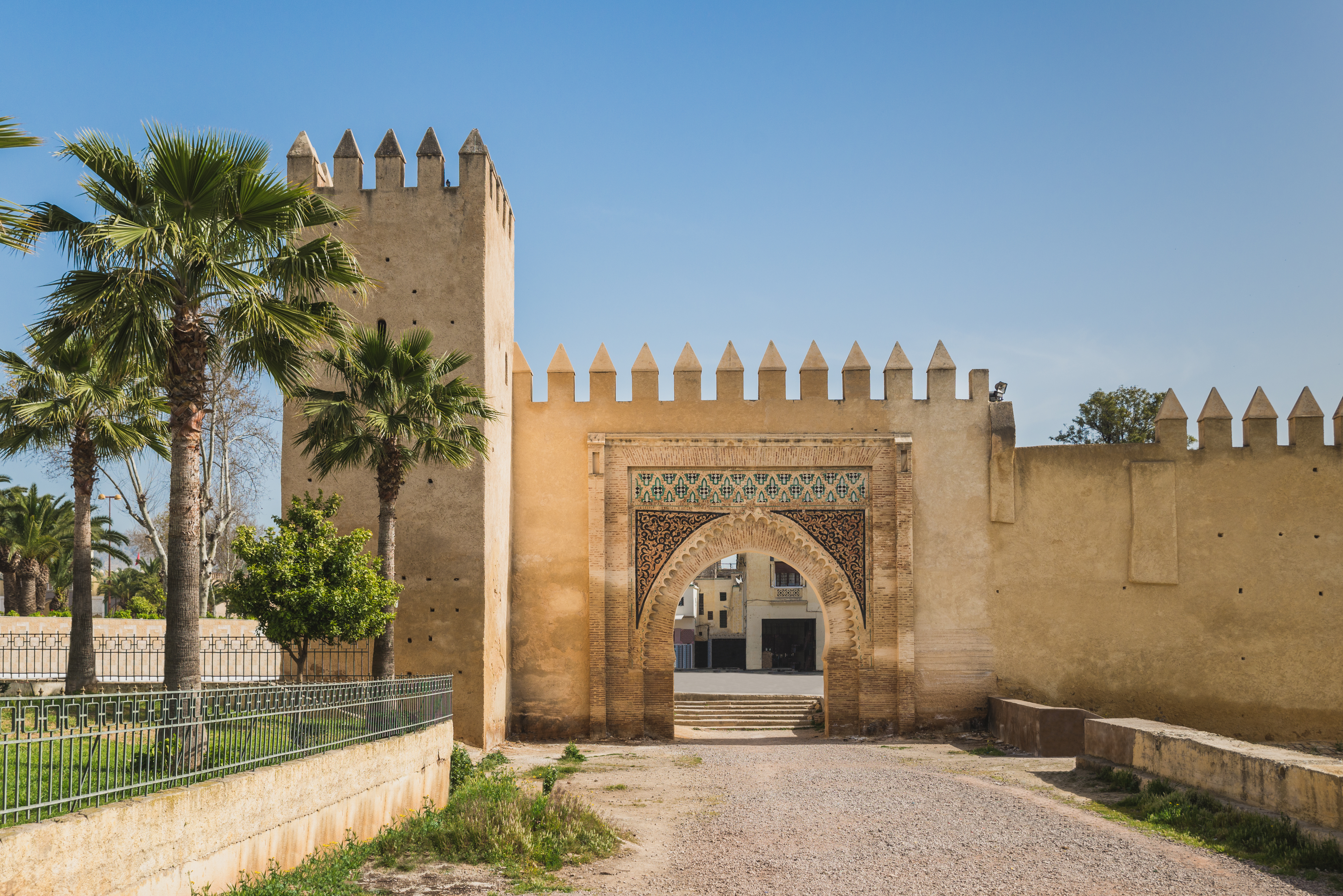 Sand-colored fortress wall with an arched gate and palm trees under a clear sky.