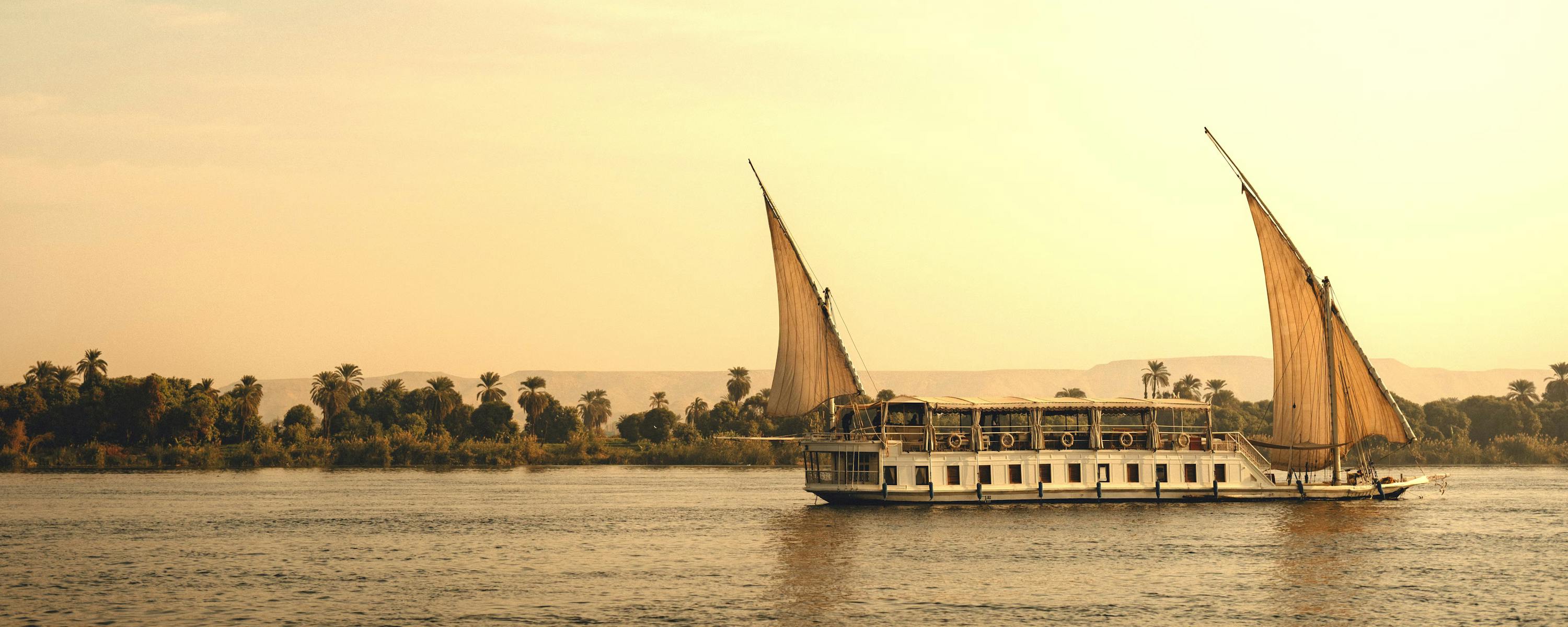 Sailboat with two white sails on a river at sunset with a palm-lined shore.