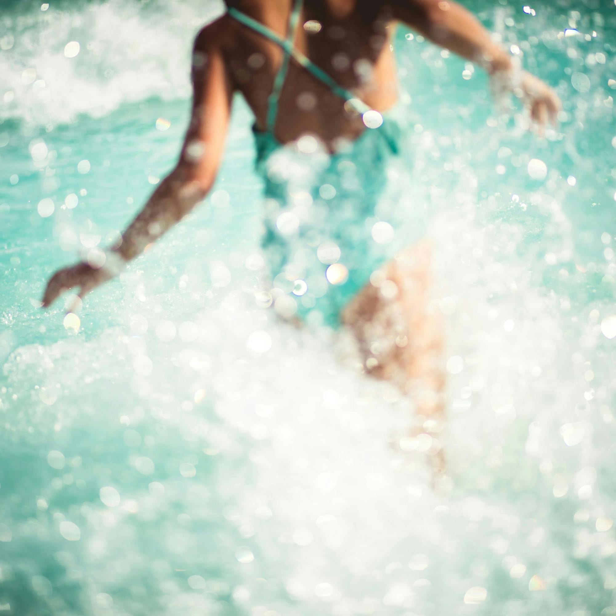 A person splashes through sunlit turquoise seawater, with bright droplets and foamy waves filling the frame.