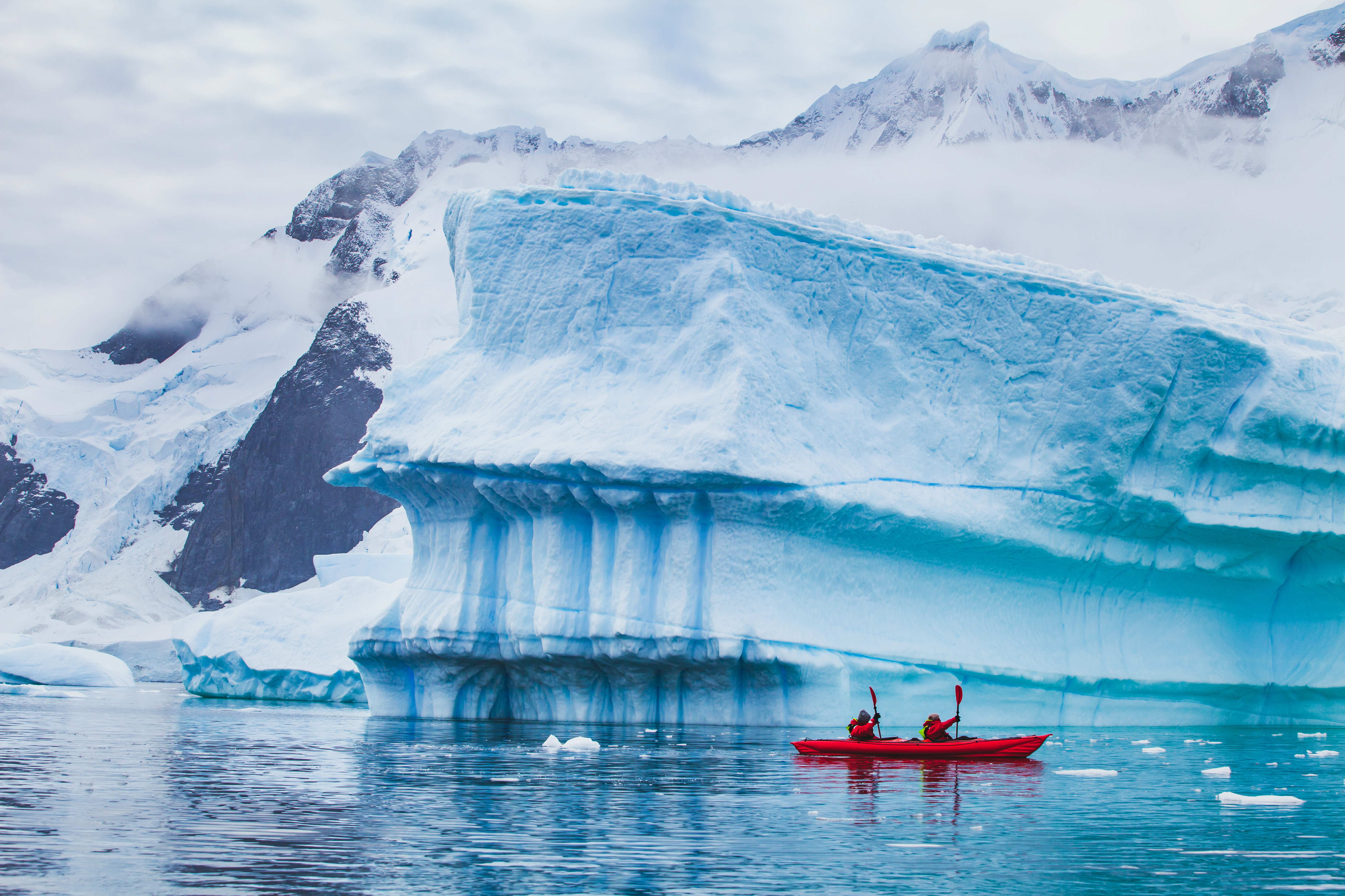A red kayak glides past a towering blue iceberg in calm water, with snowy mountains and scattered ice nearby.