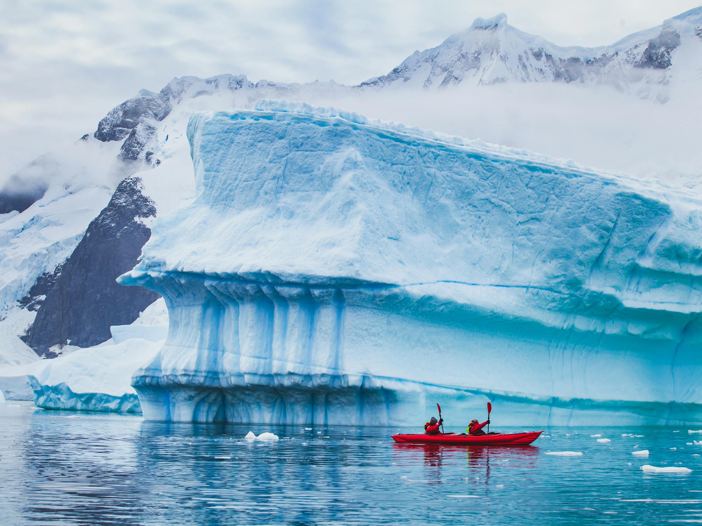 A red kayak glides past a towering blue iceberg in calm water, with snowy mountains and scattered ice nearby.