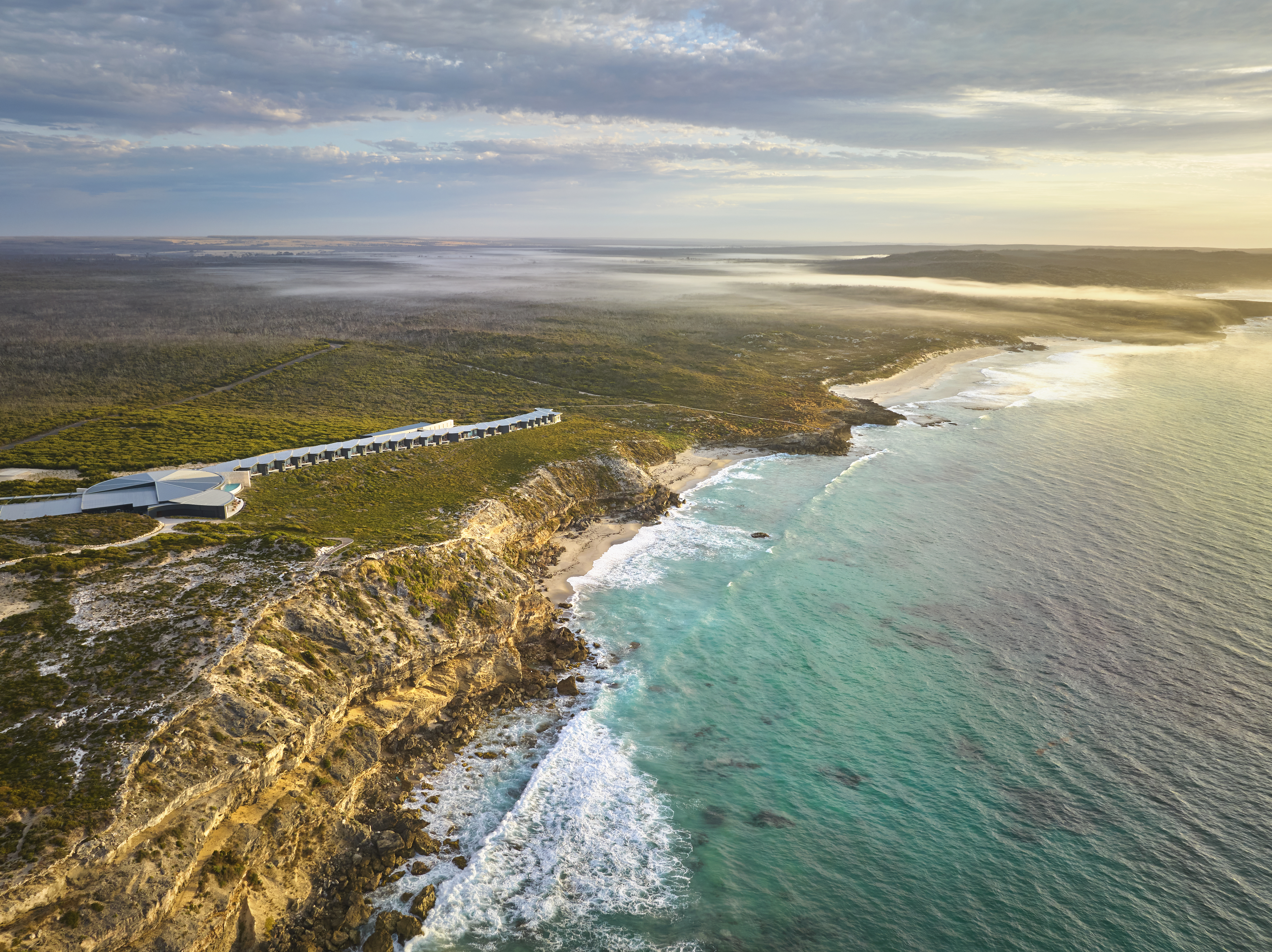 Aerial view of a lodge on rugged sea cliffs, with waves breaking below and turquoise water stretching to the horizon.