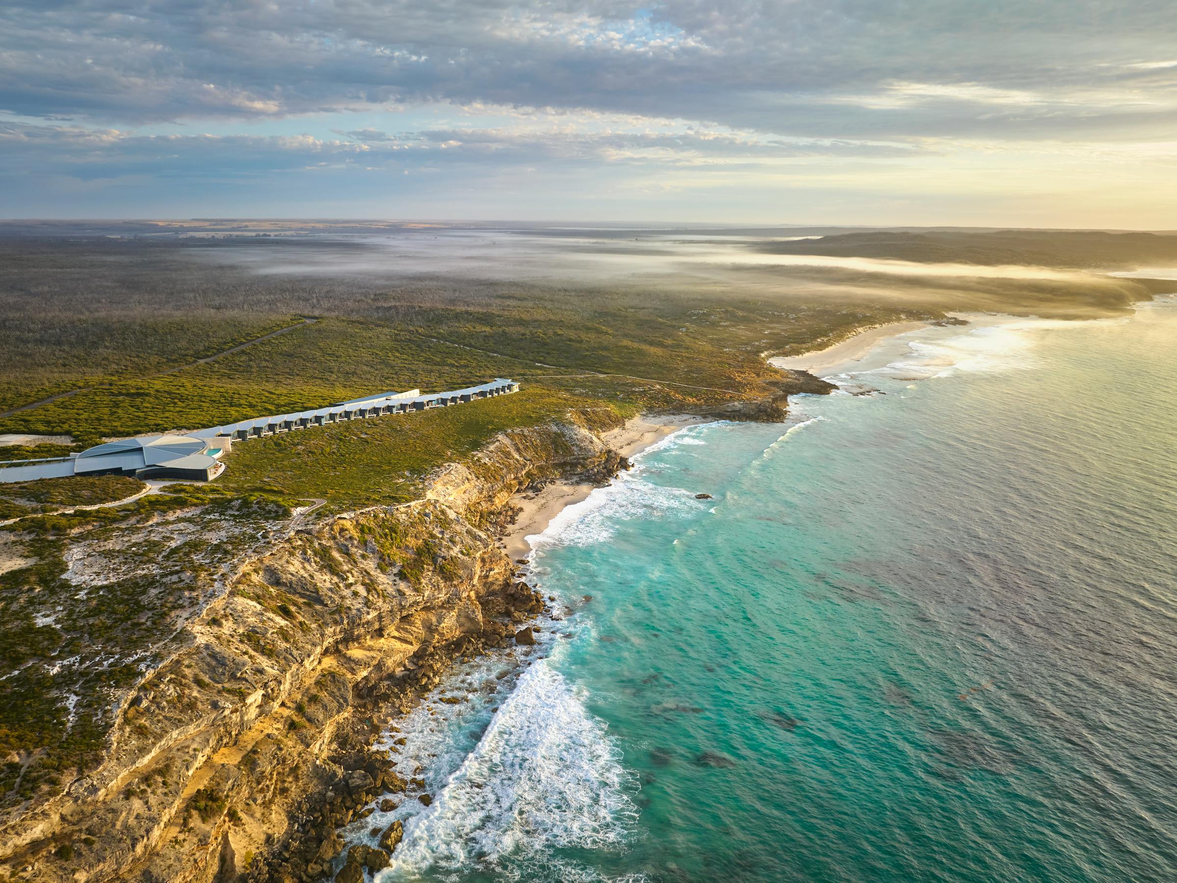 Aerial view of a lodge on rugged sea cliffs, with waves breaking below and turquoise water stretching to the horizon.