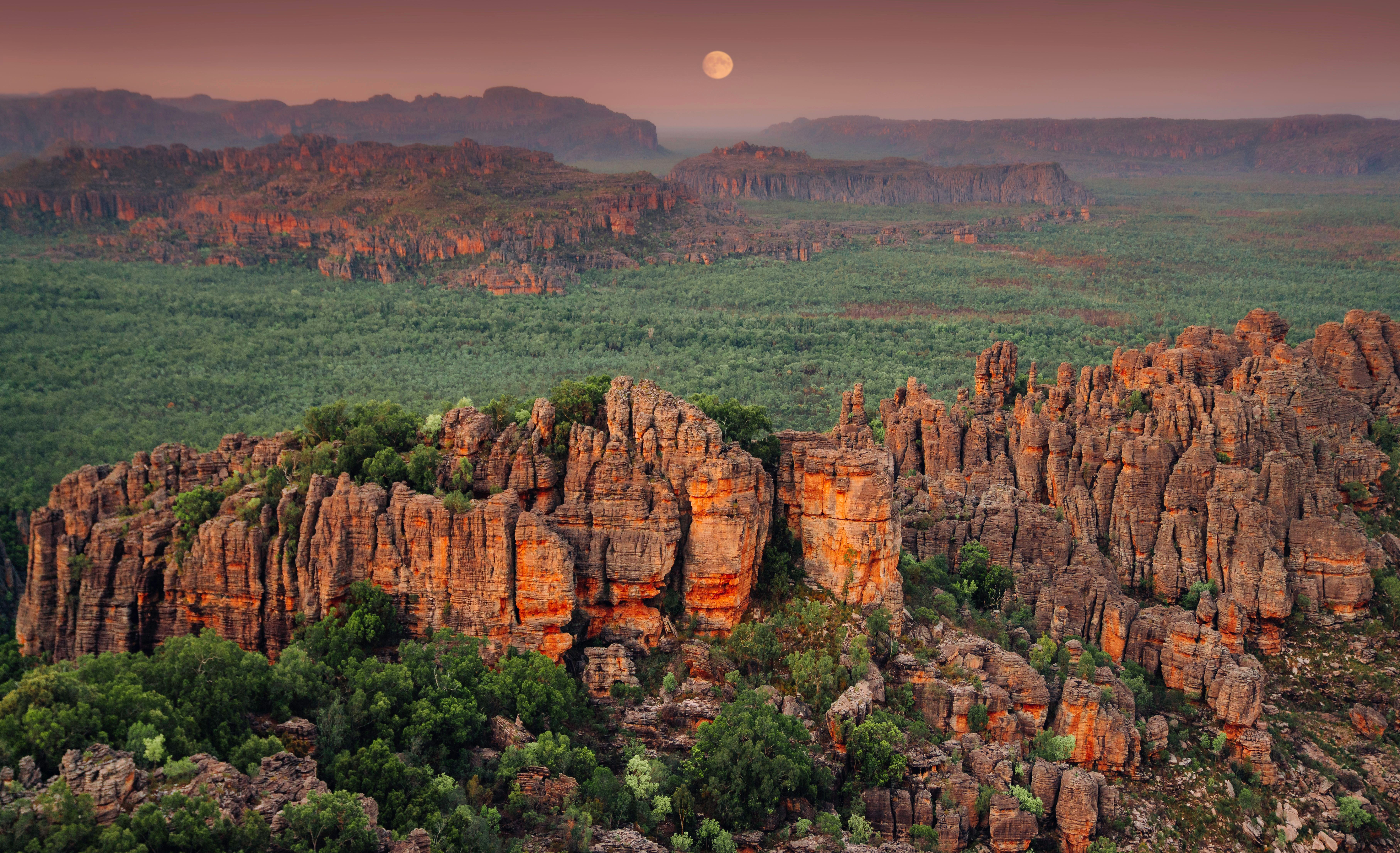 Red sandstone escarpment rises above green woodland at dusk, with a full moon hanging over distant ridges.