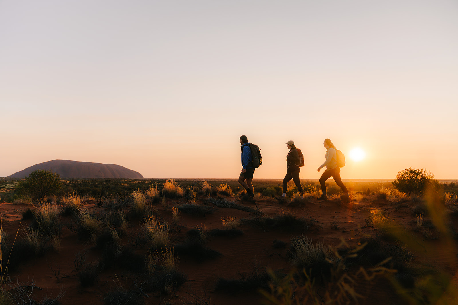 Silhouetted hikers climb a sandy ridge at sunset, with a deep orange sky and low desert shrubs below them.