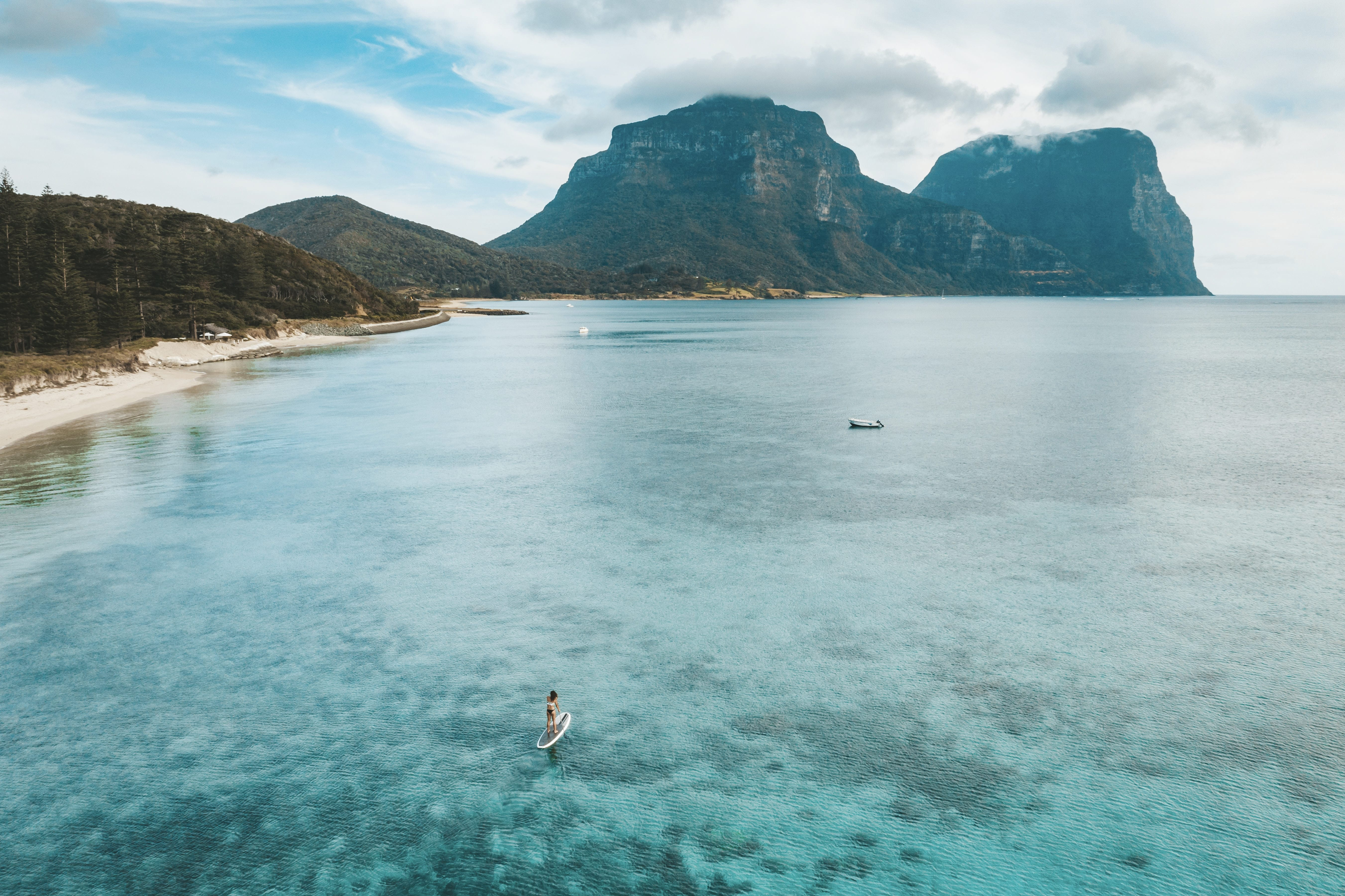 Quiet sandy beach meets a turquoise lagoon as steep green peaks rise beyond the shoreline beneath soft morning light.