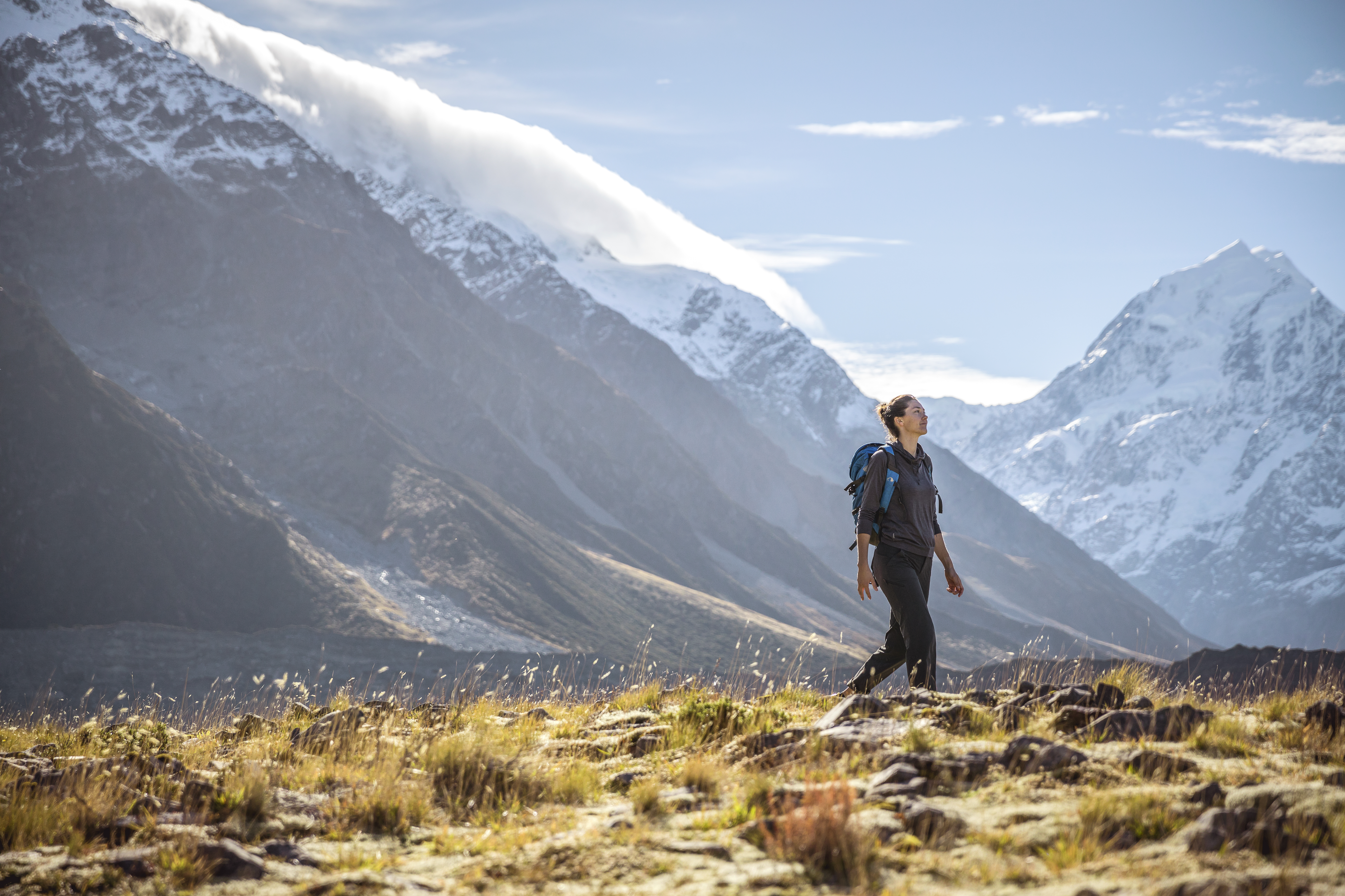 Hiker stands on a grassy ridge facing snowy peaks, with a wide glacier valley stretching into the distance.