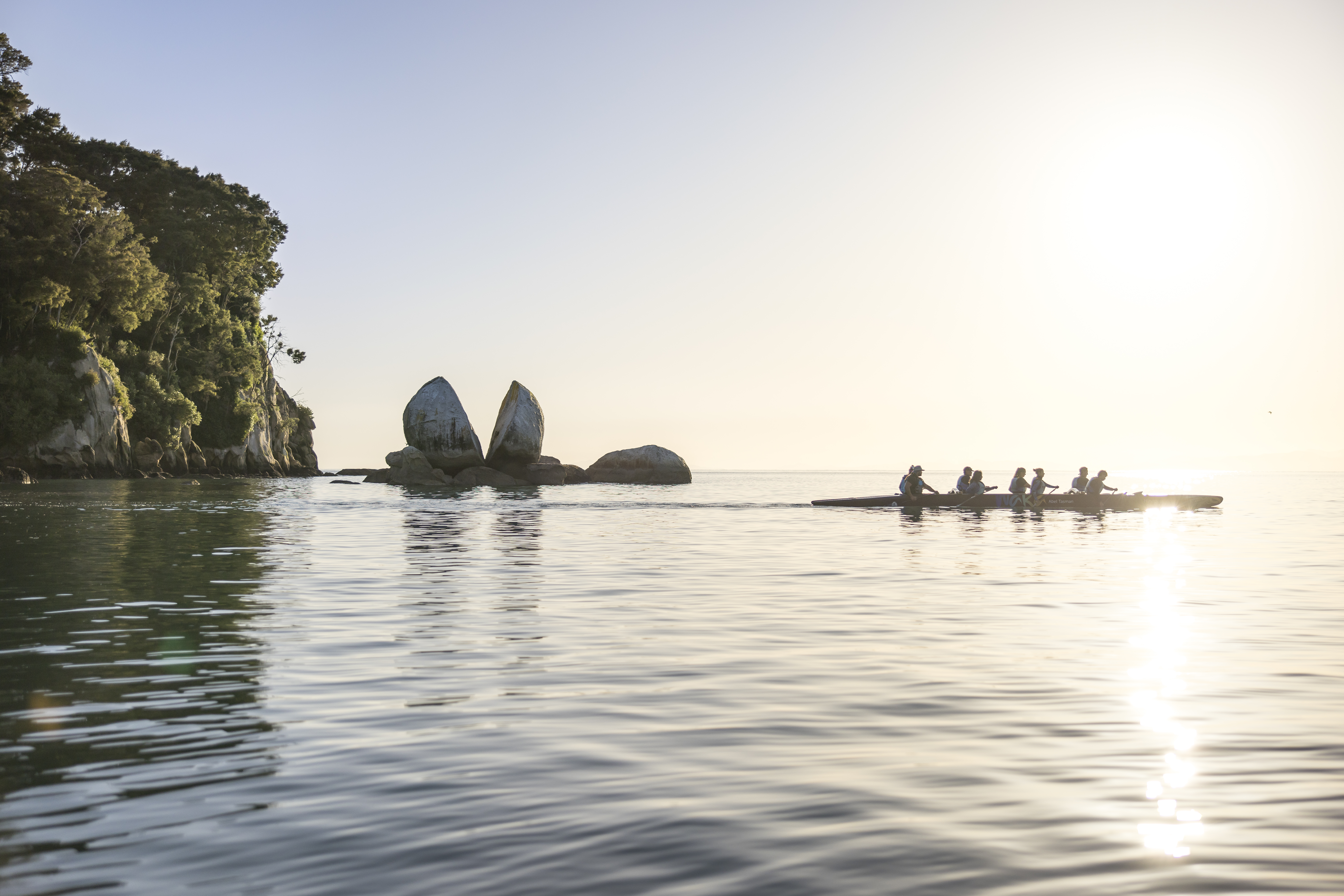 Kayakers paddle across calm water toward rocky islets at sunrise, with golden light reflecting across the bay.