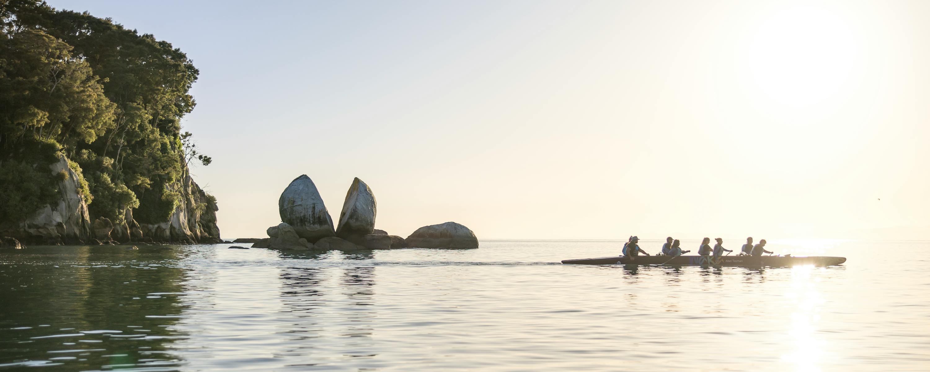Kayakers paddle across calm water toward rocky islets at sunrise, with golden light reflecting across the bay.