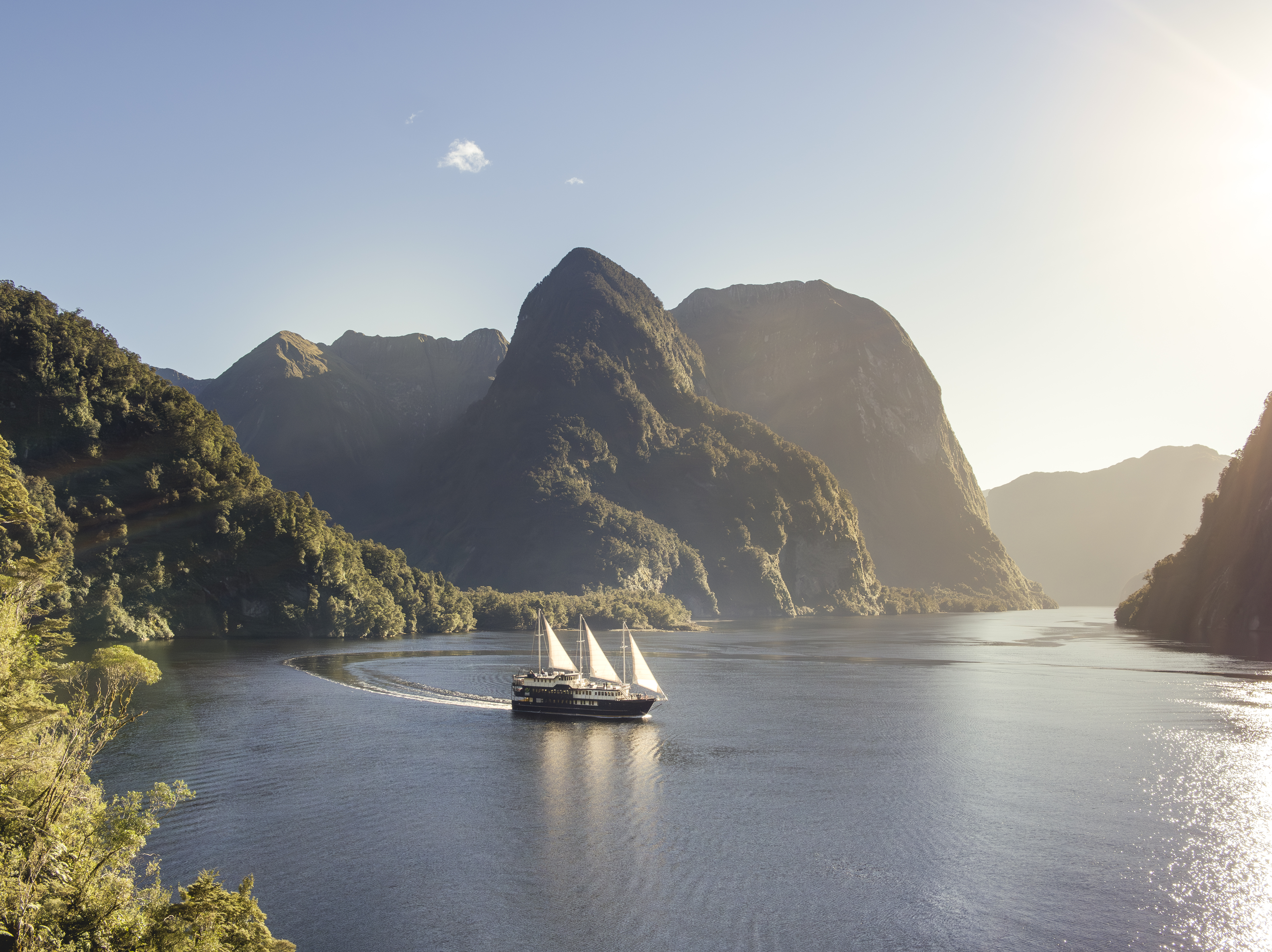 White sailboat glides through a steep-sided fjord in bright sun, with sheer mountains and calm water all around.