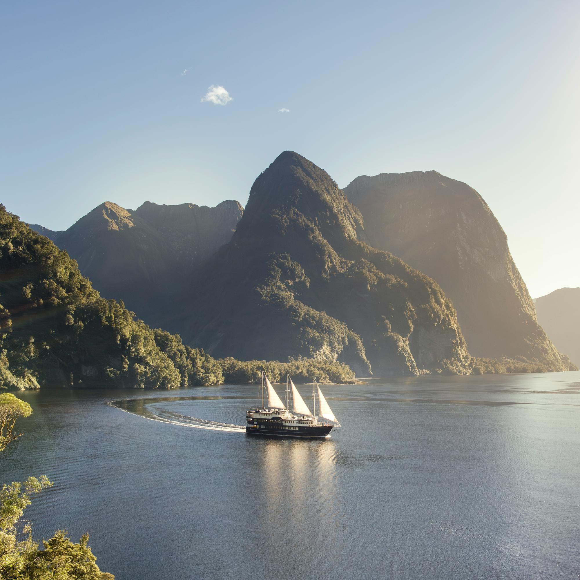 White sailboat glides through a steep-sided fjord in bright sun, with sheer mountains and calm water all around.