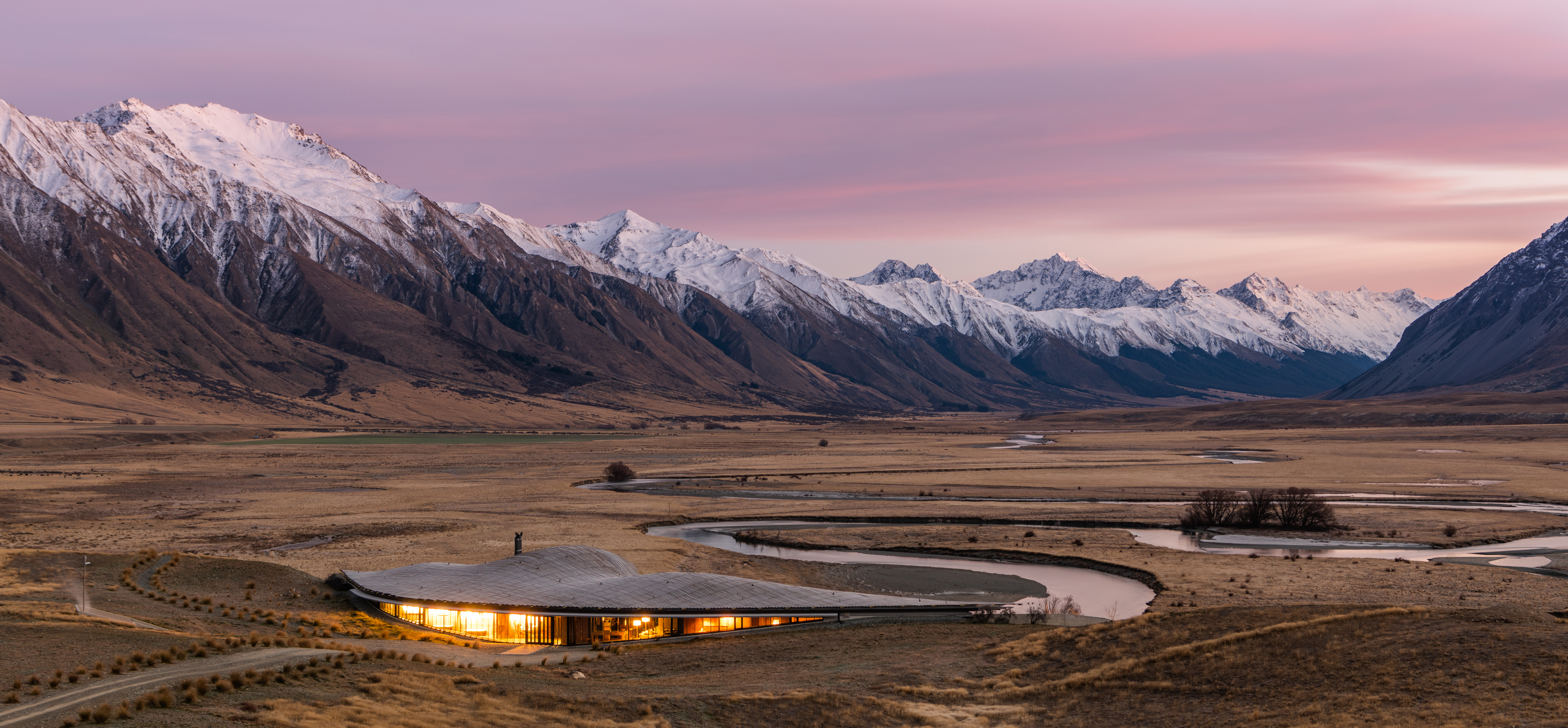 Modern lodge glows at dusk beside a winding river, framed by golden plains and snowcapped mountains beyond.