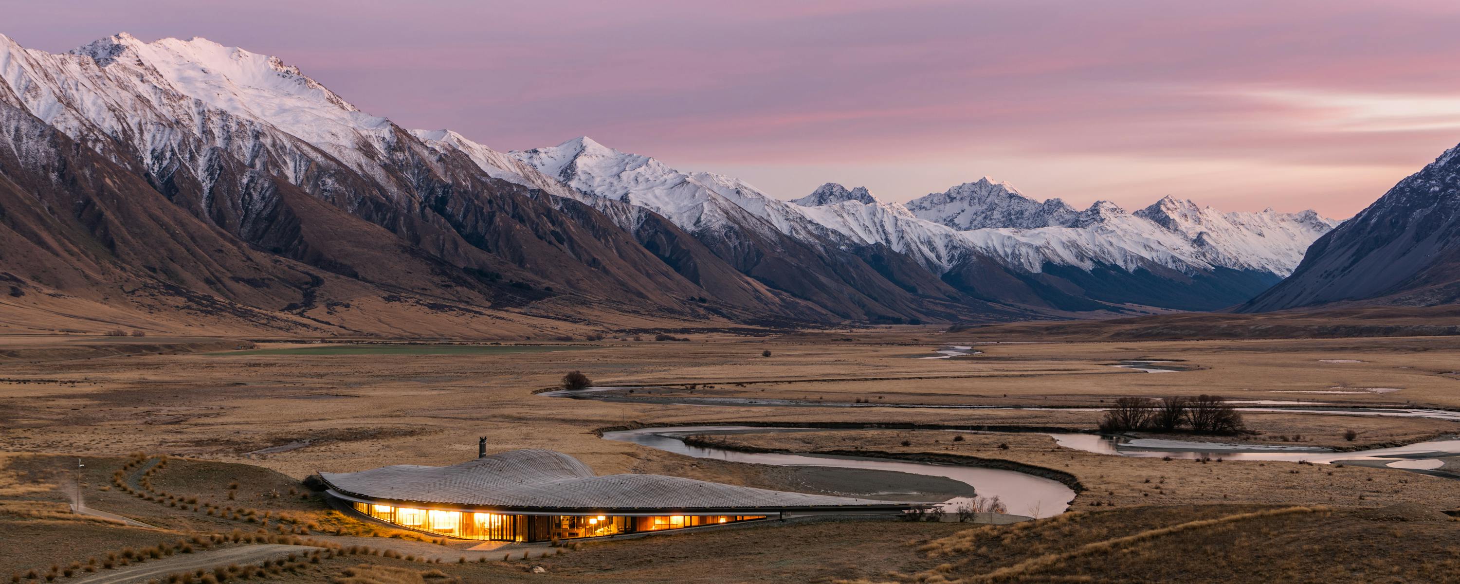 Modern lodge glows at dusk beside a winding river, framed by golden plains and snowcapped mountains beyond.