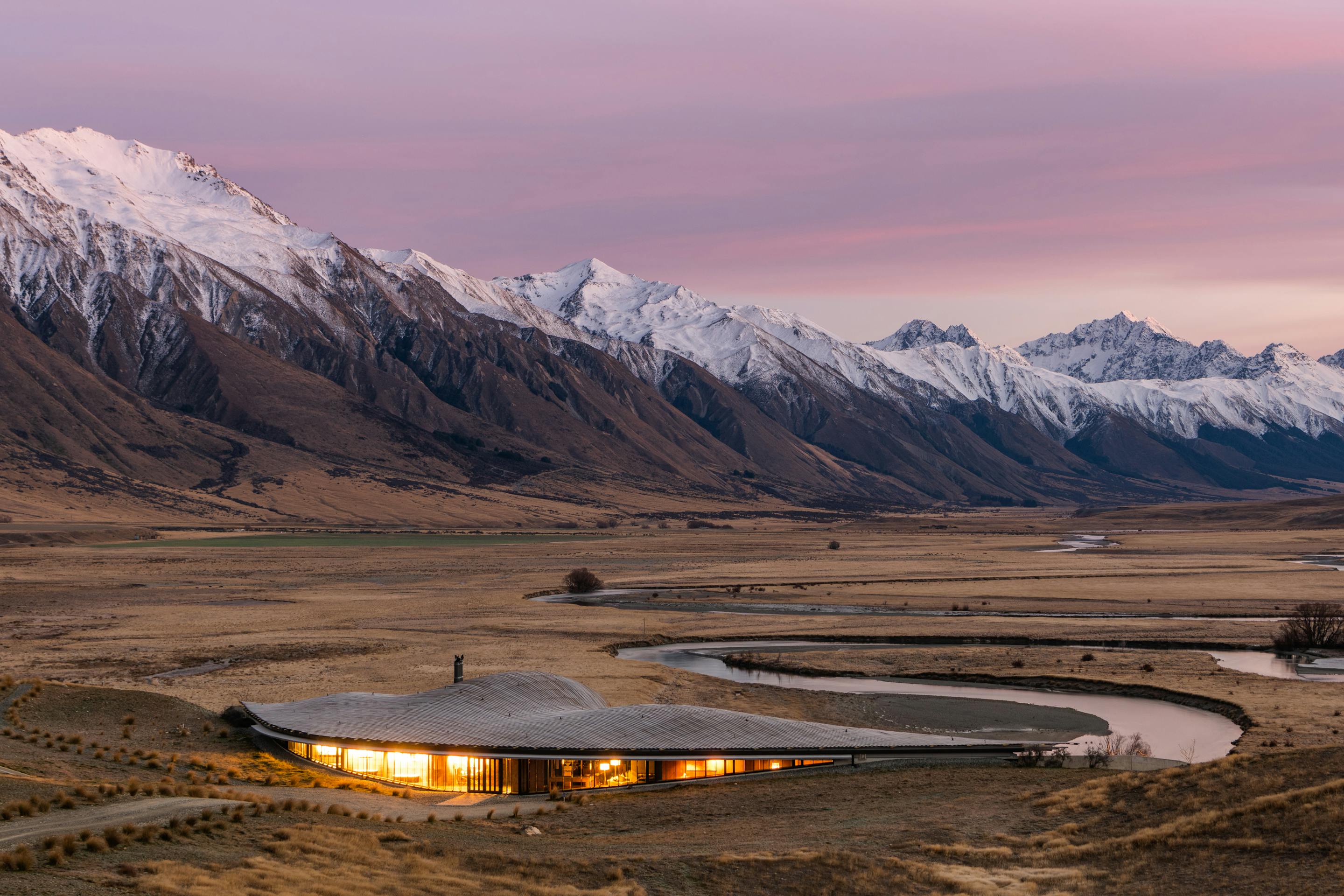 Modern lodge glows at dusk beside a winding river, framed by golden plains and snowcapped mountains beyond.