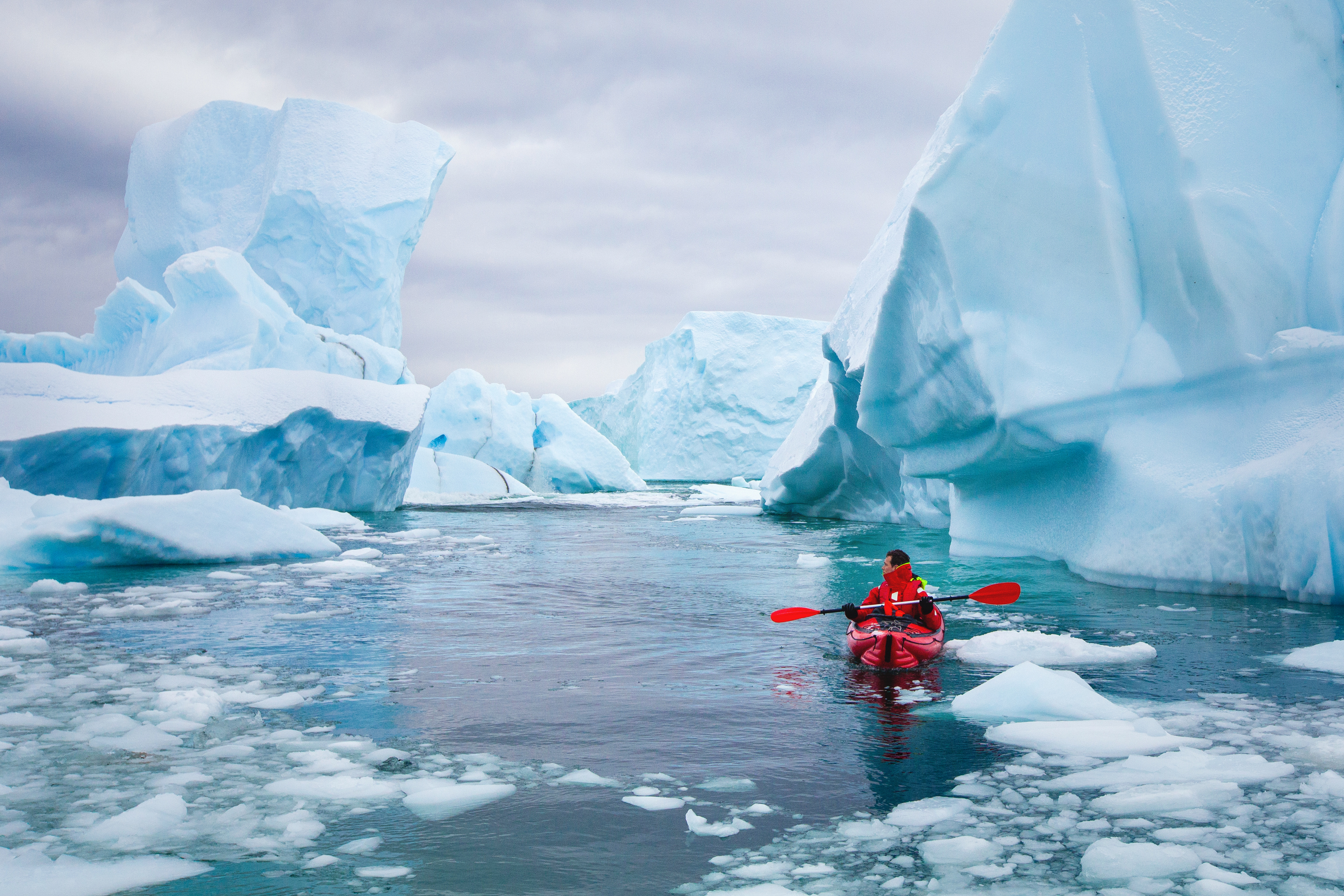 A kayaker paddles through a channel of blue icebergs on glassy water, with snow-covered peaks beyond the bay.