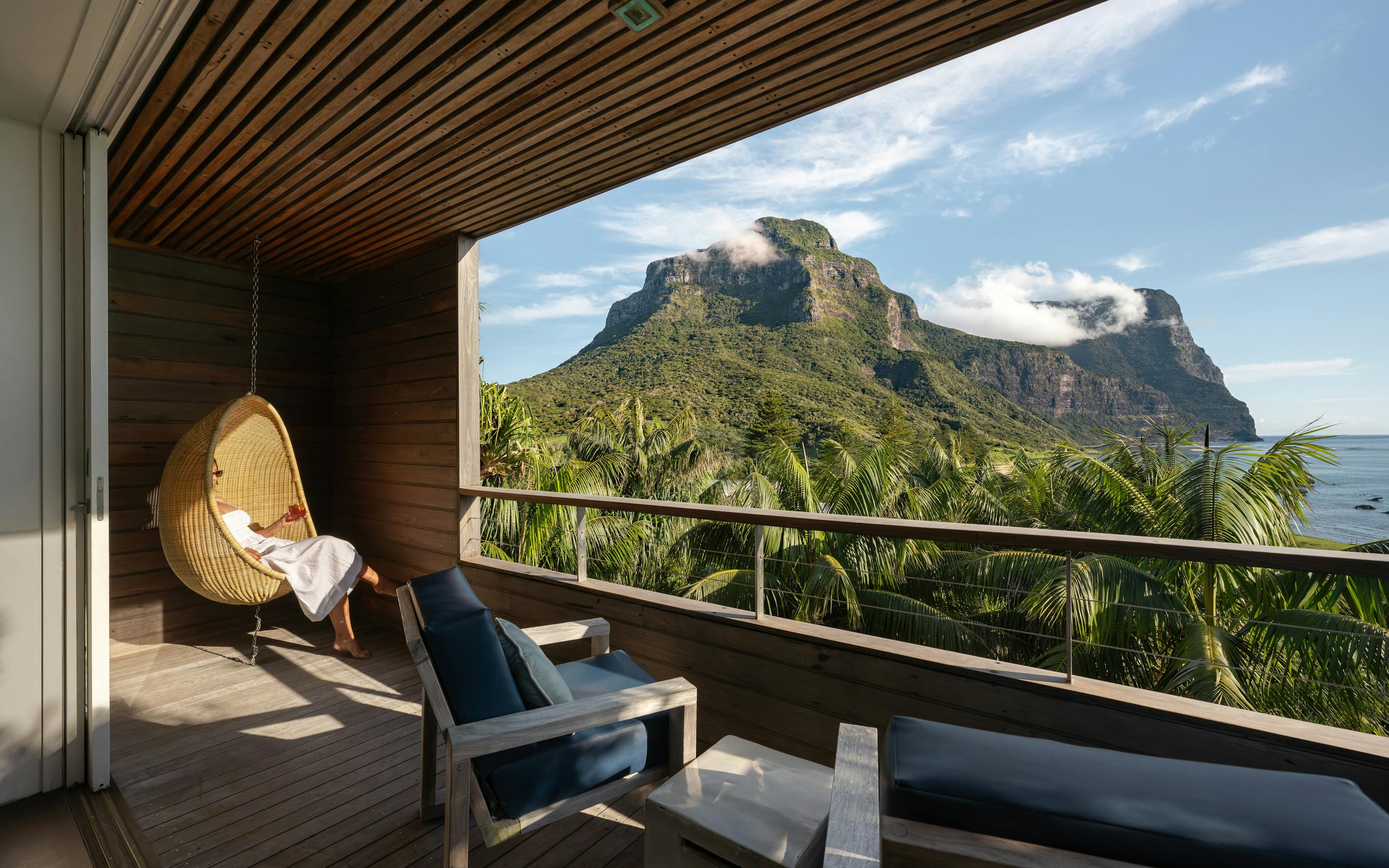 Covered lodge deck with lounge chairs and a daybed overlooks lush palms and rugged mountains beneath a cloudy sky.