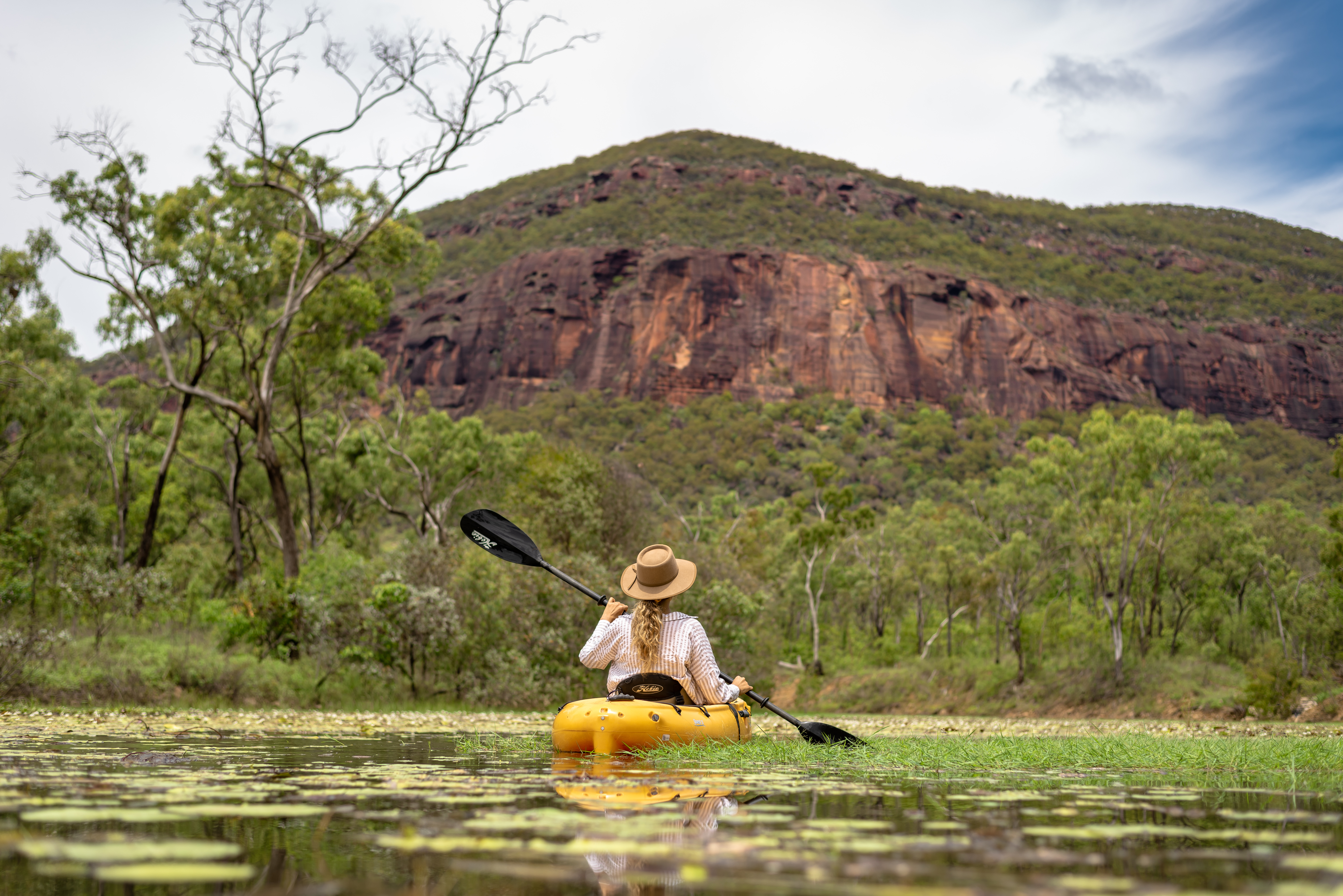 Person paddles a kayak across a calm lagoon dotted with lily pads, with a rugged rock massif rising behind.
