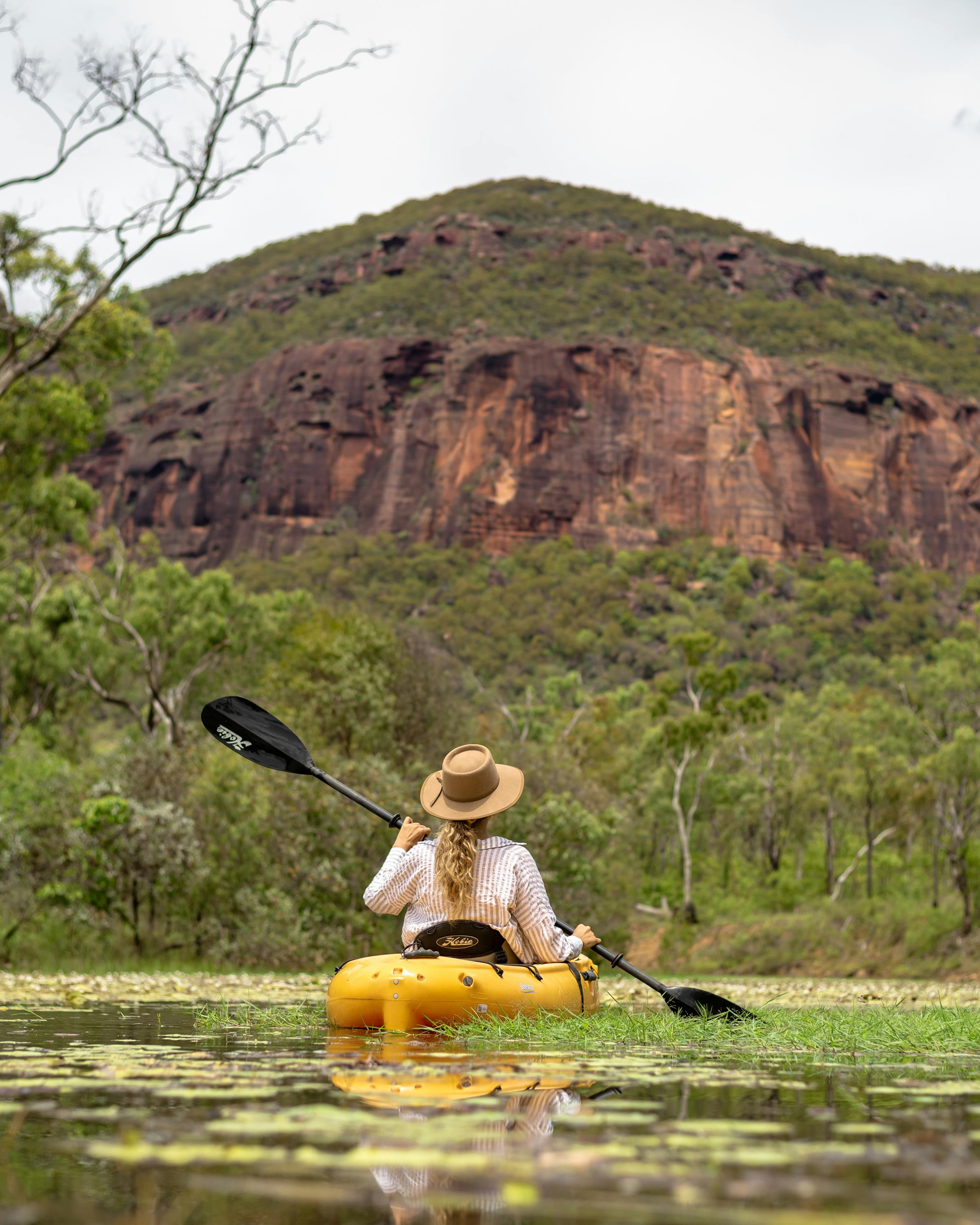 Person paddles a kayak across a calm lagoon dotted with lily pads, with a rugged rock massif rising behind.