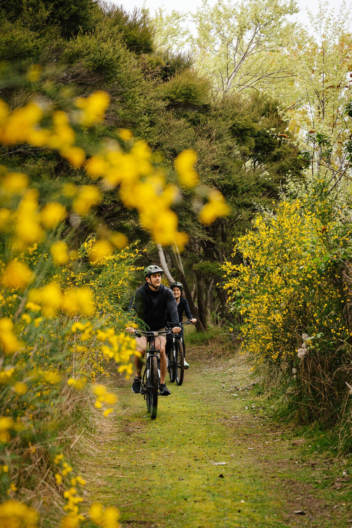 Cyclist rides along a narrow green path bordered by yellow flowers and shrubs, with bright sunlight ahead.