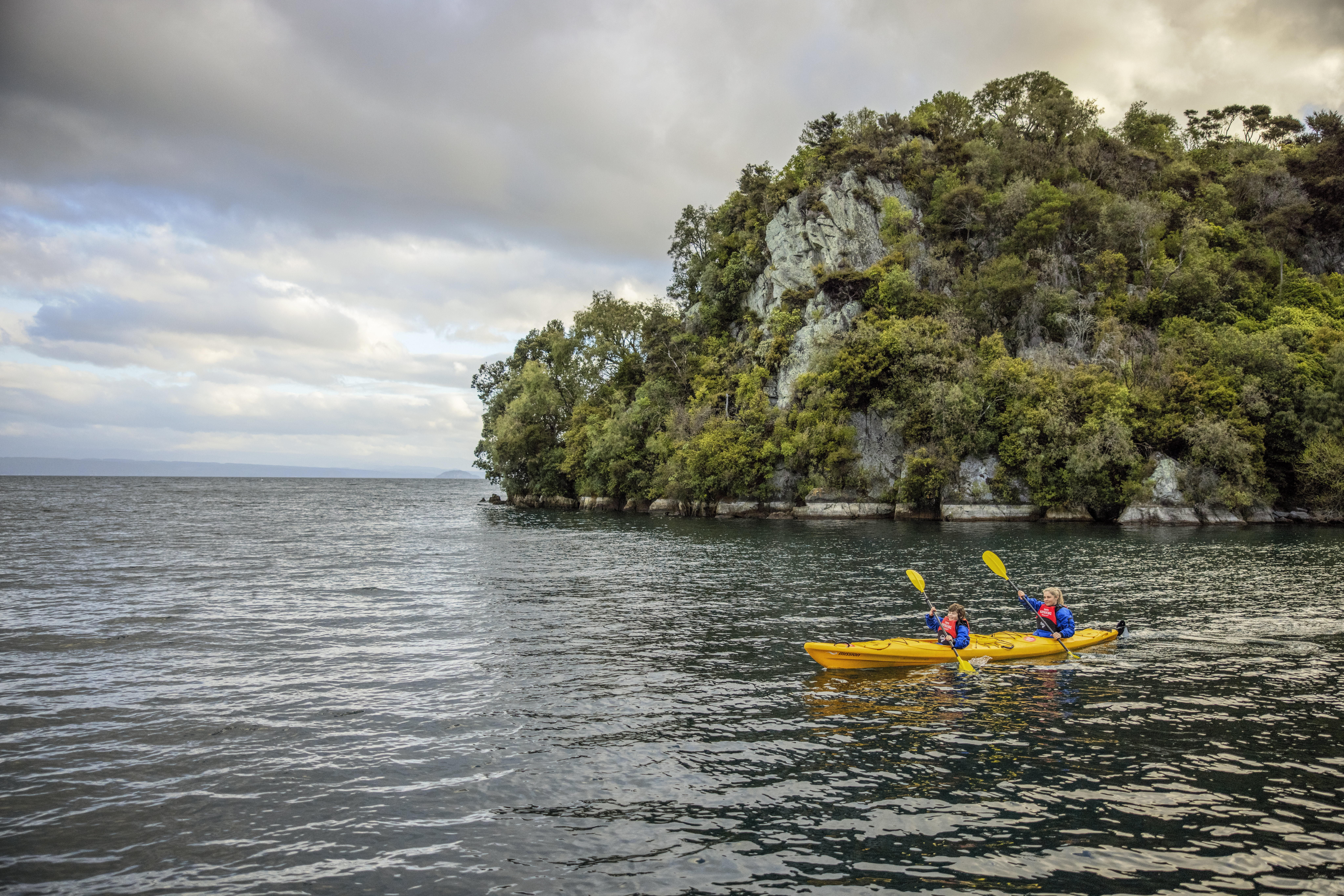 Two people paddle bright kayaks on calm water near a wooded island, with gentle ripples under an overcast sky.