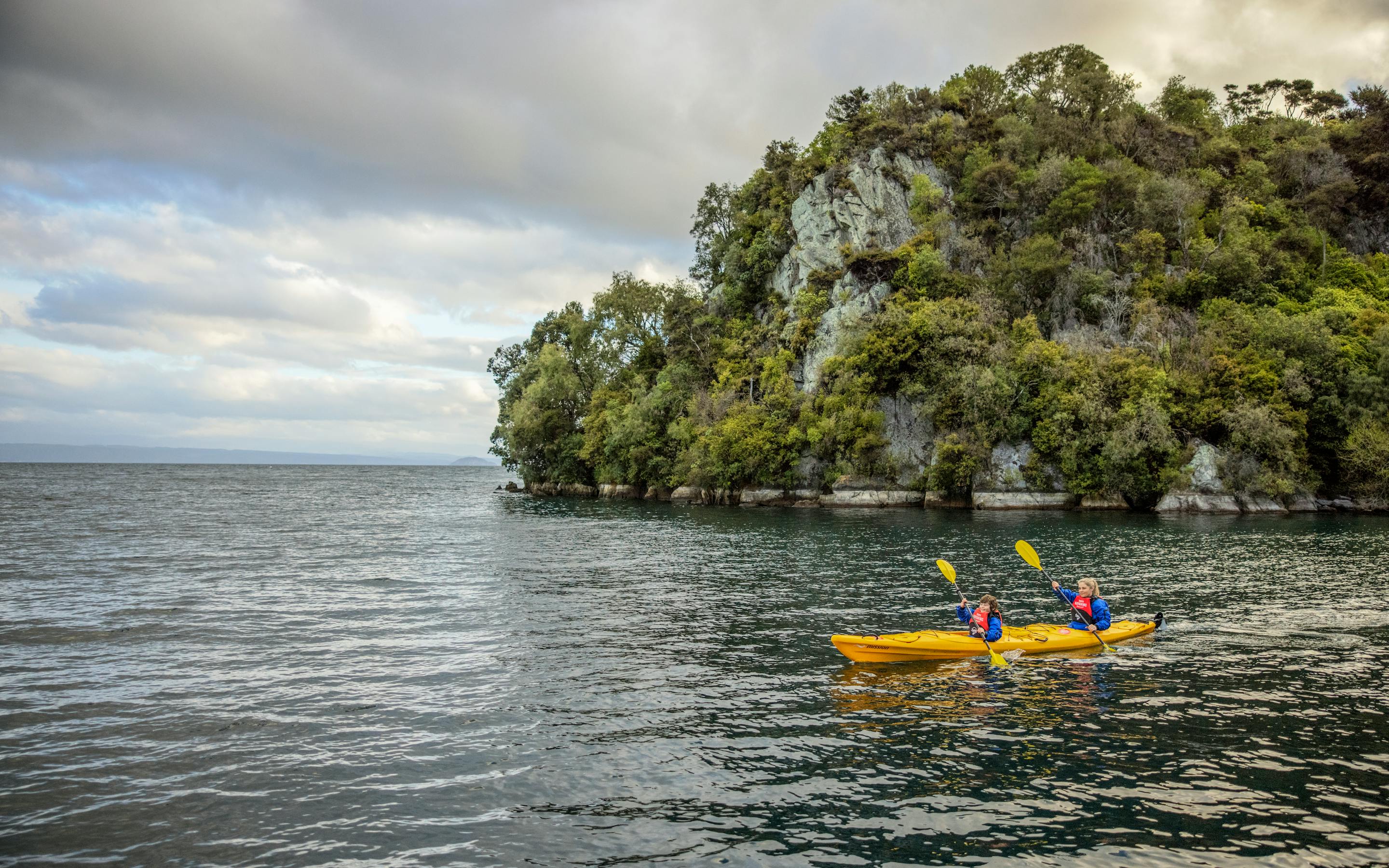 Two people paddle bright kayaks on calm water near a wooded island, with gentle ripples under an overcast sky.