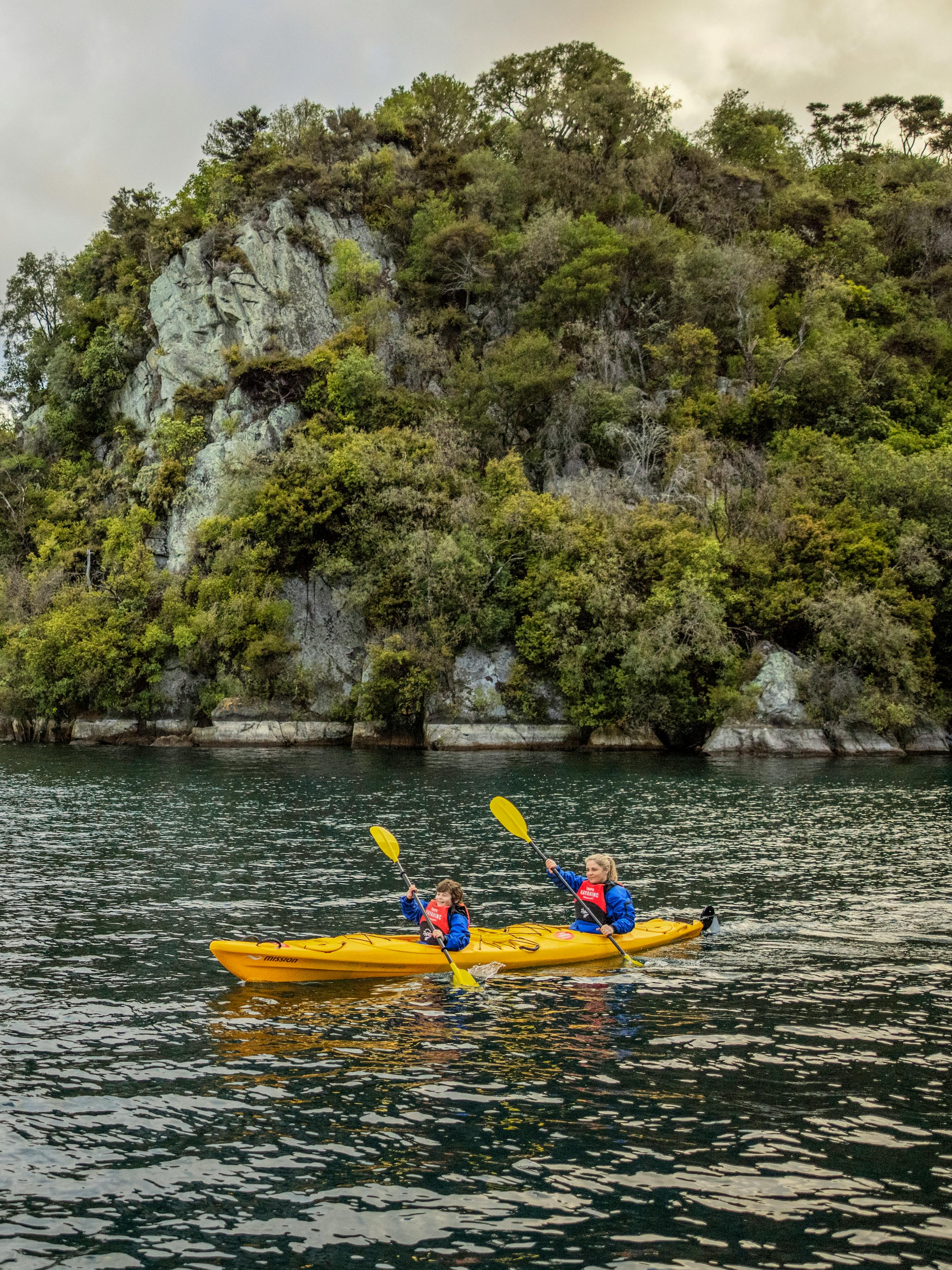 Two people paddle bright kayaks on calm water near a wooded island, with gentle ripples under an overcast sky.