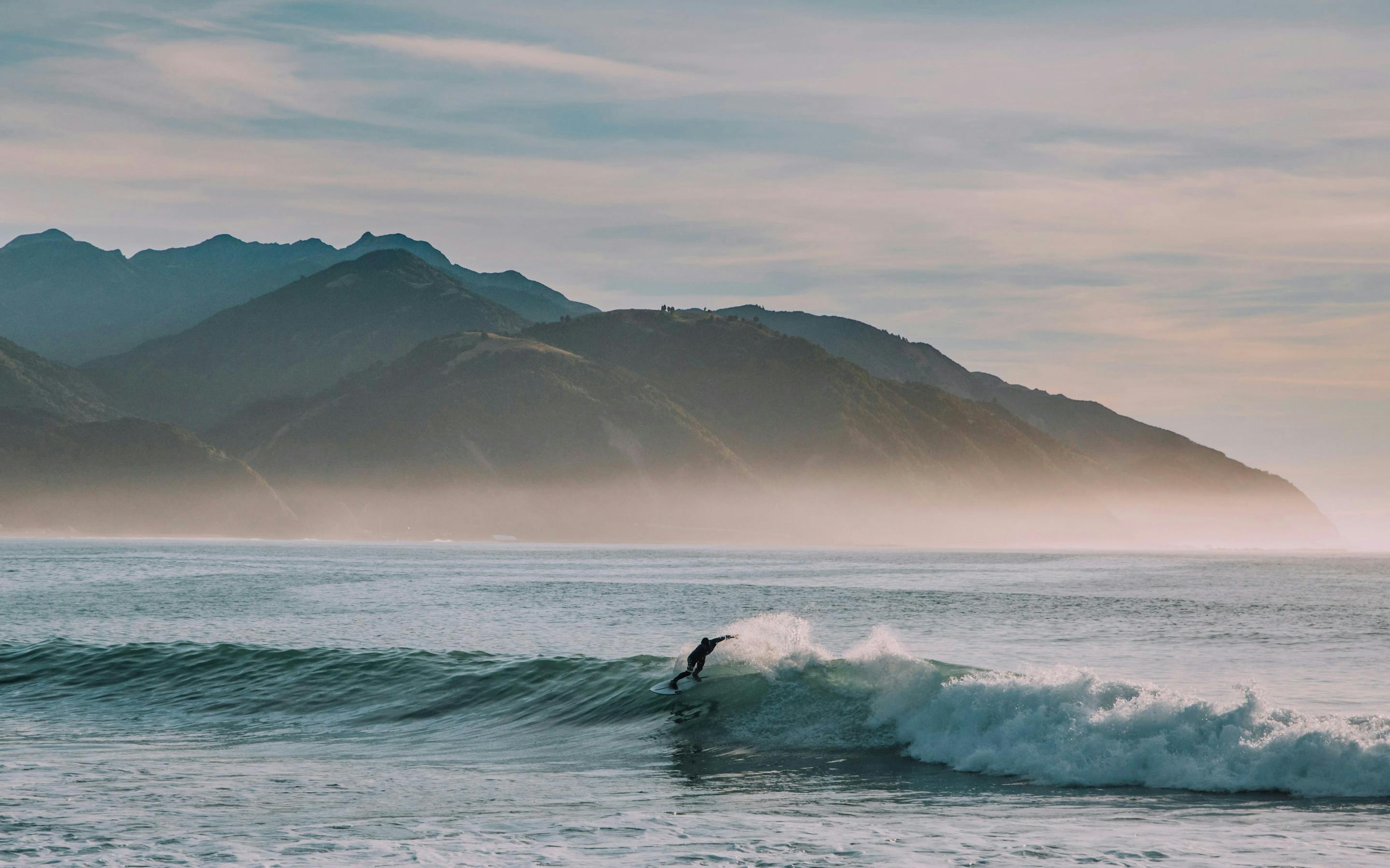 Surfer rides a breaking wave near shore, with misty mountains behind and pale pink sunrise light on the horizon.