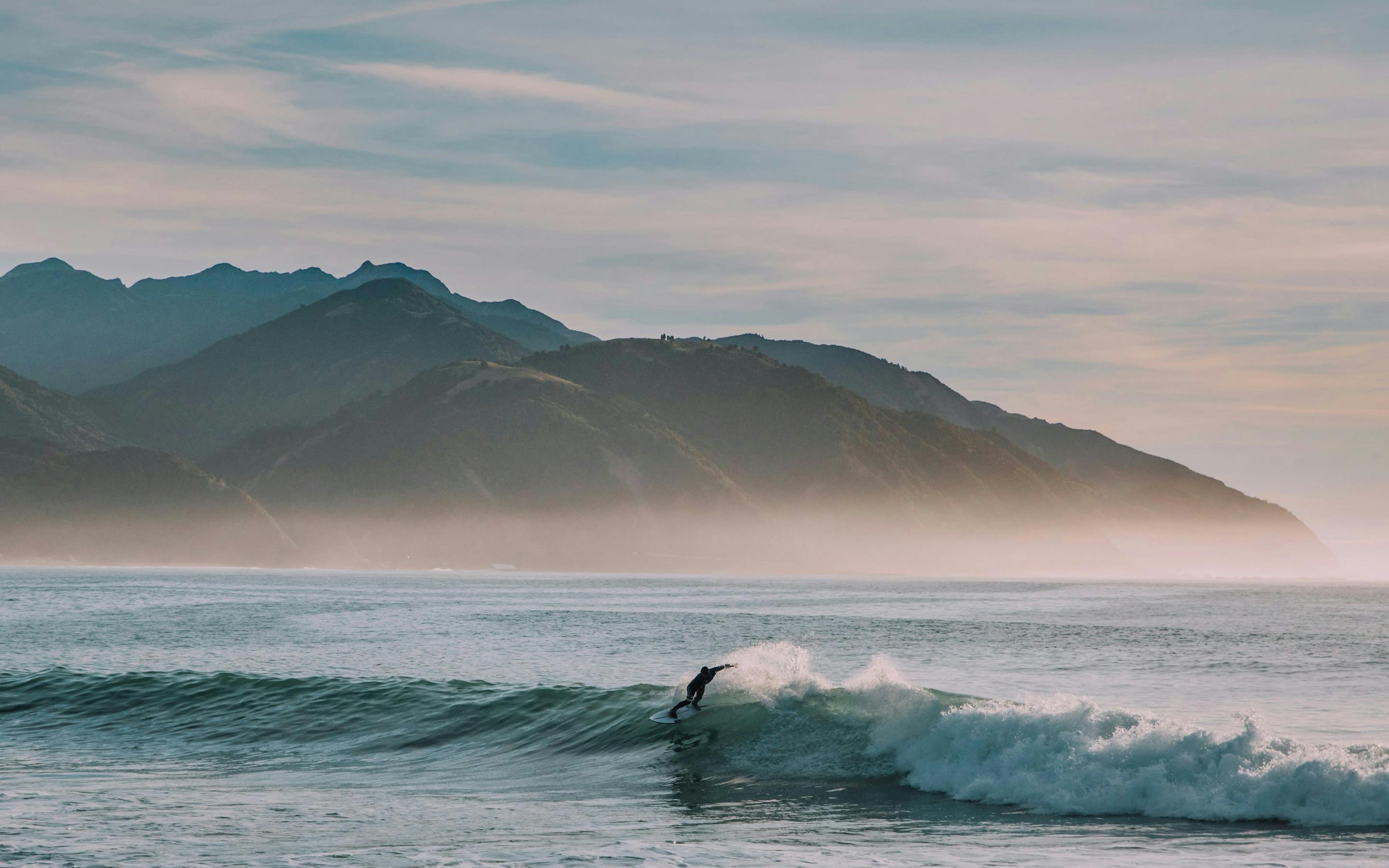 Surfer rides a breaking wave near shore, with misty mountains behind and pale pink sunrise light on the horizon.