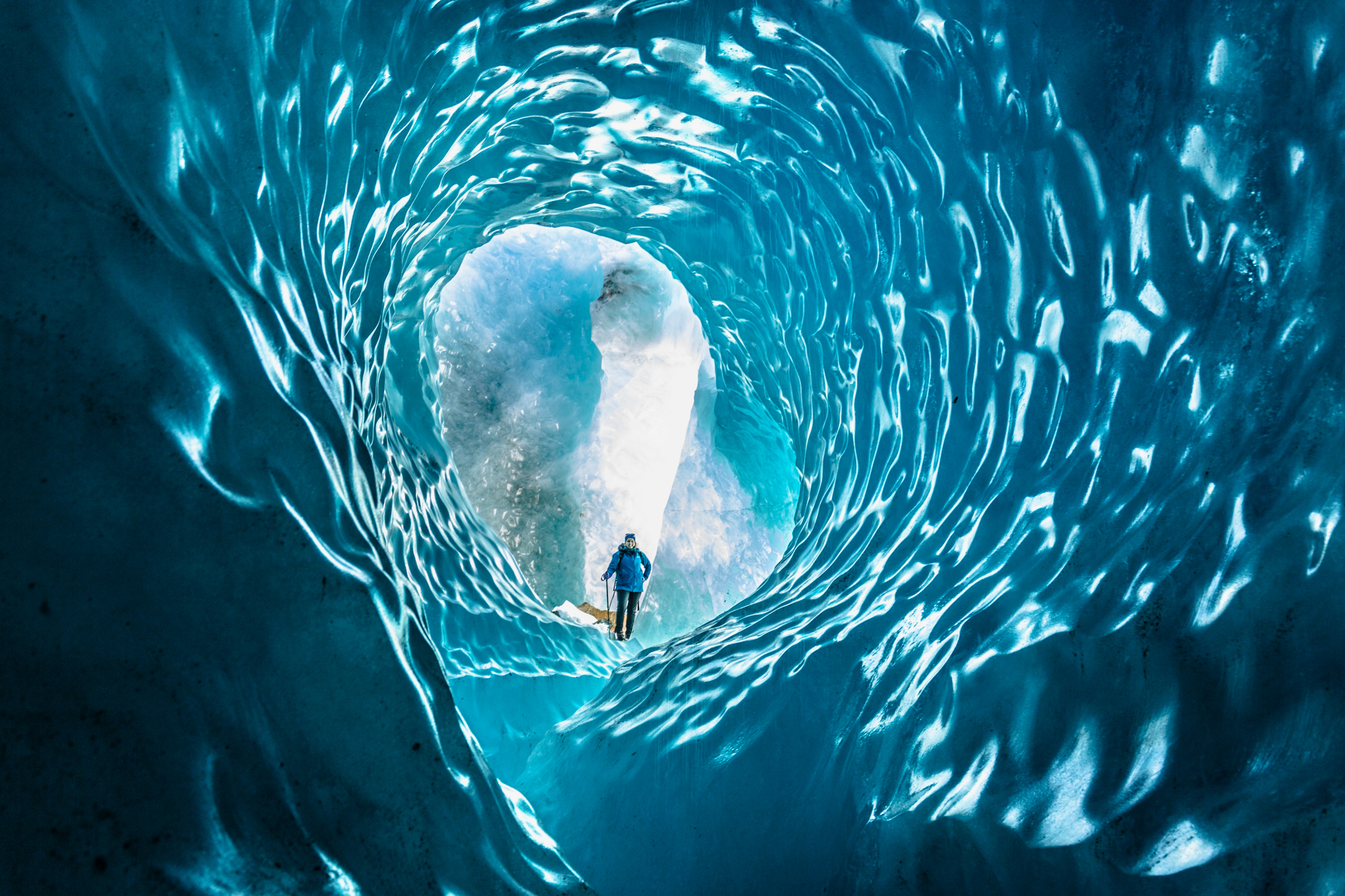 Person stands inside a blue ice cave tunnel, framed by swirling textures as light spills in from the entrance.