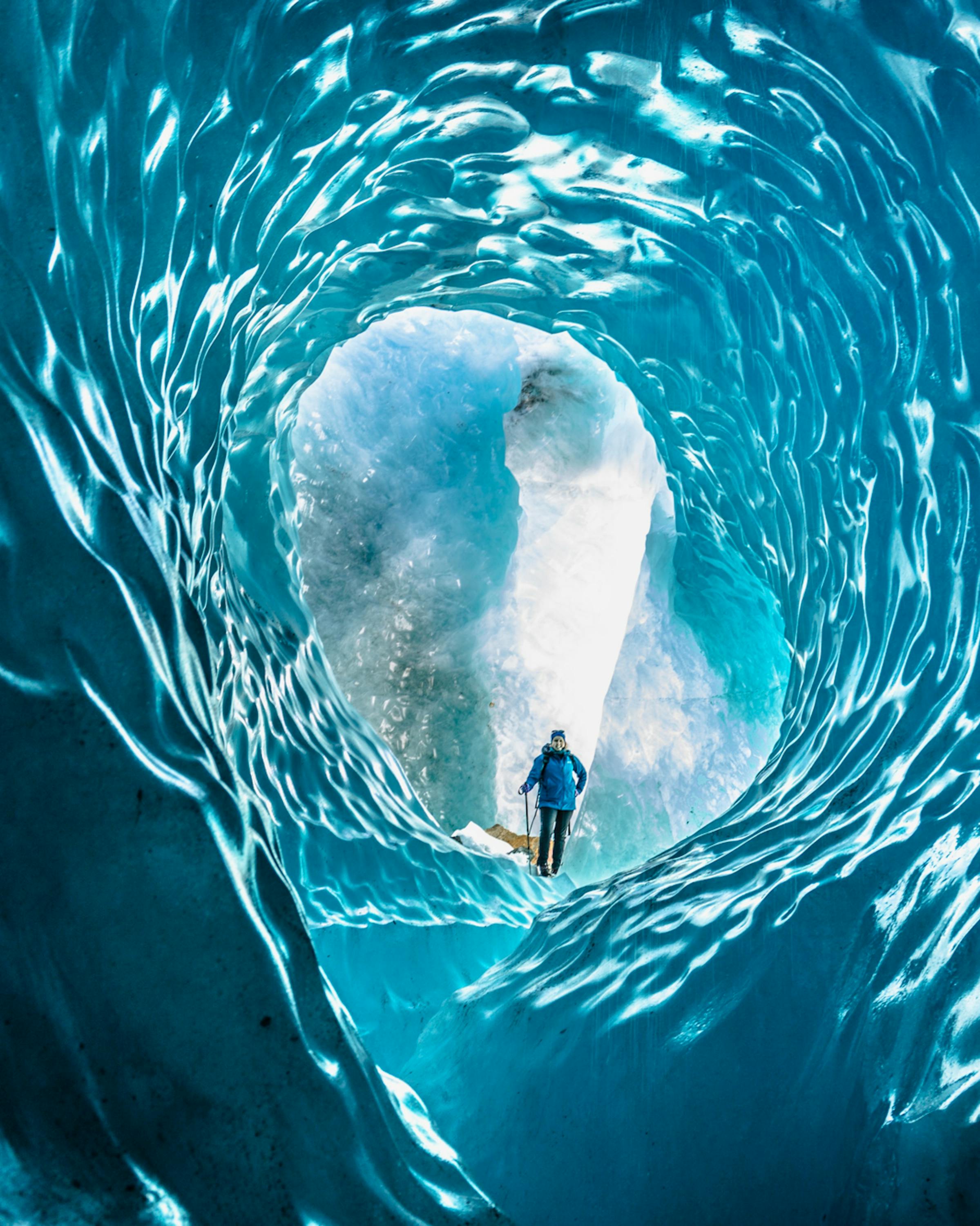 Person stands inside a blue ice cave tunnel, framed by swirling textures as light spills in from the entrance.