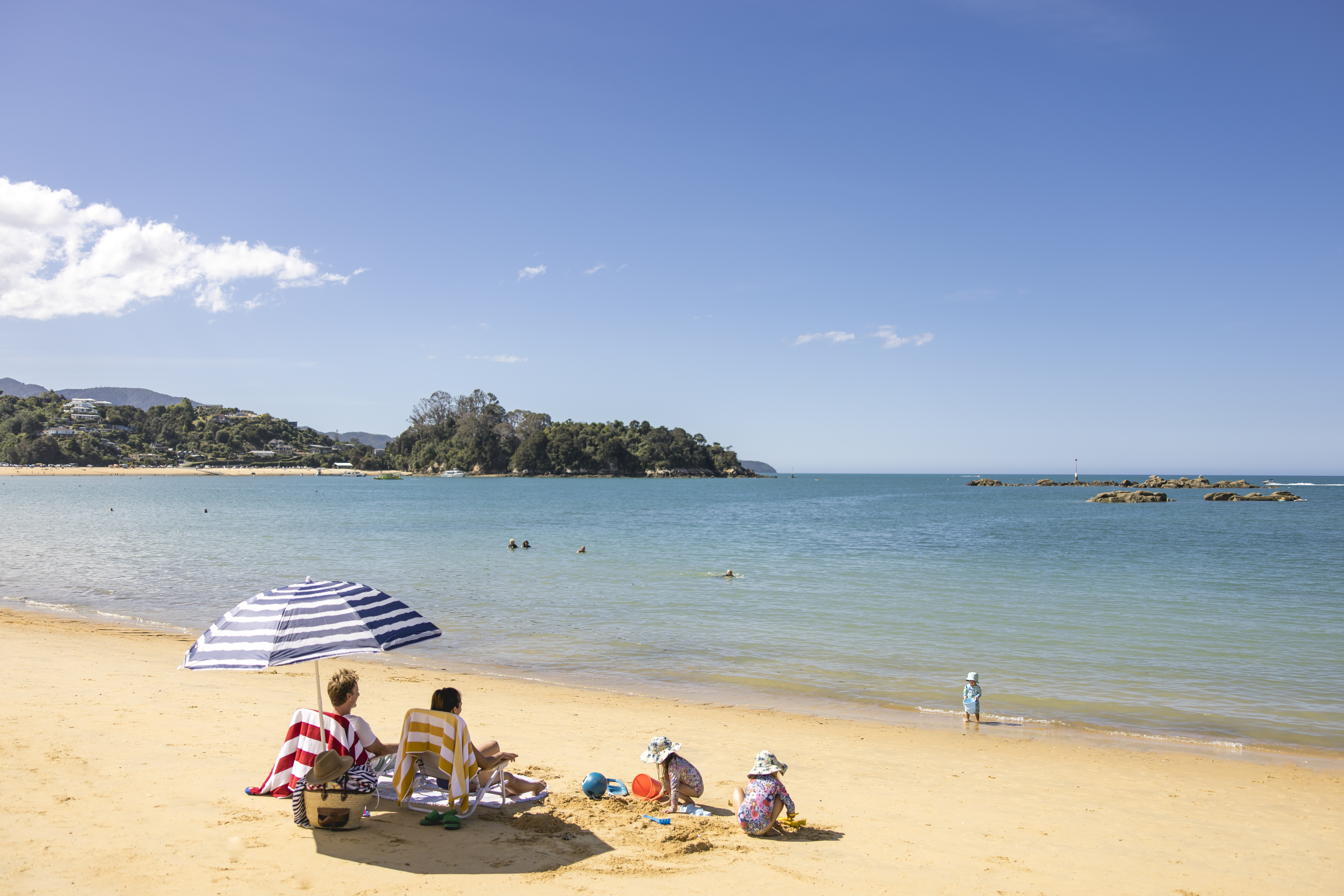 Families relax on a sandy beach beside calm turquoise water, with a striped umbrella and swimmers offshore.
