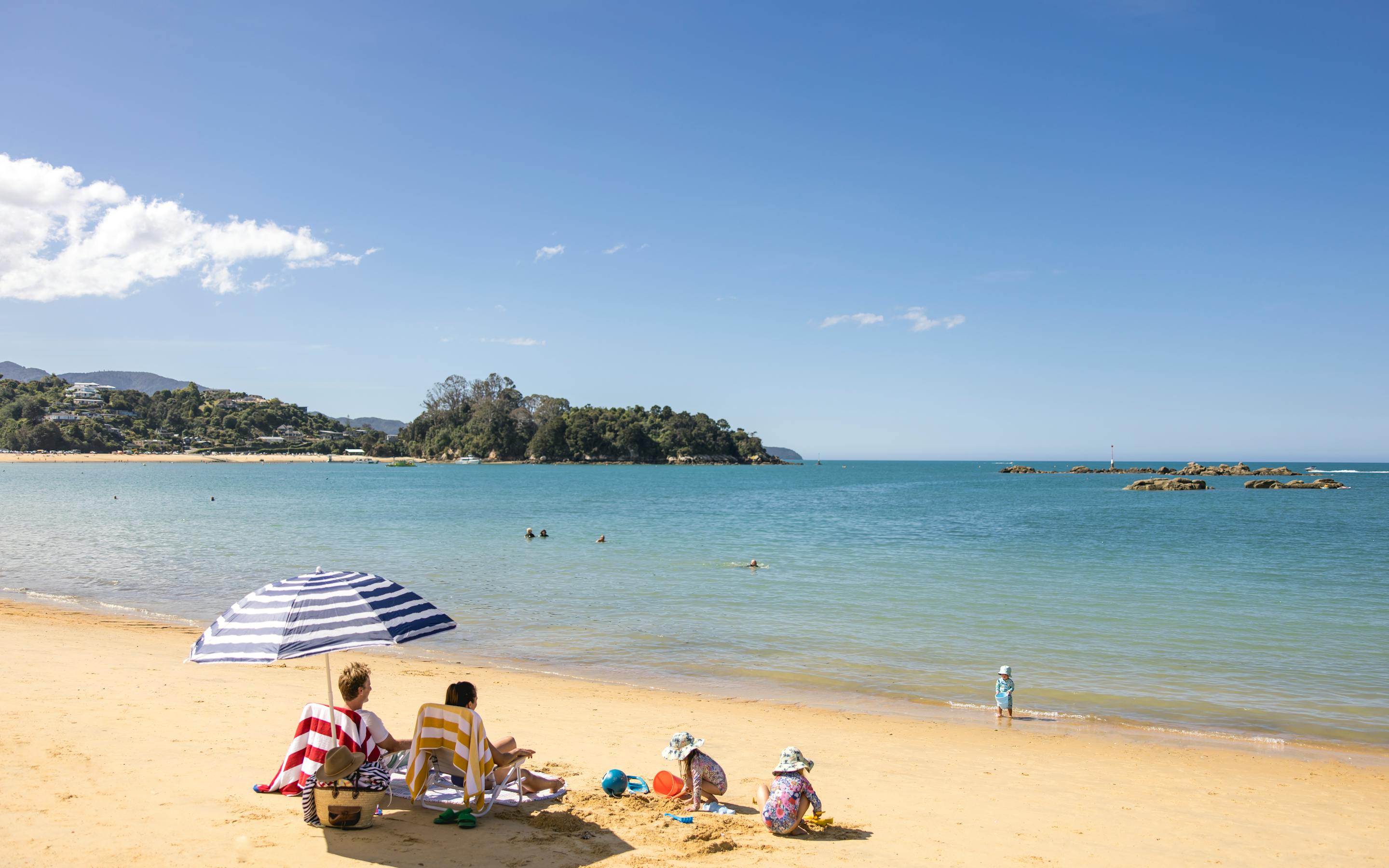 Families relax on a sandy beach beside calm turquoise water, with a striped umbrella and swimmers offshore.