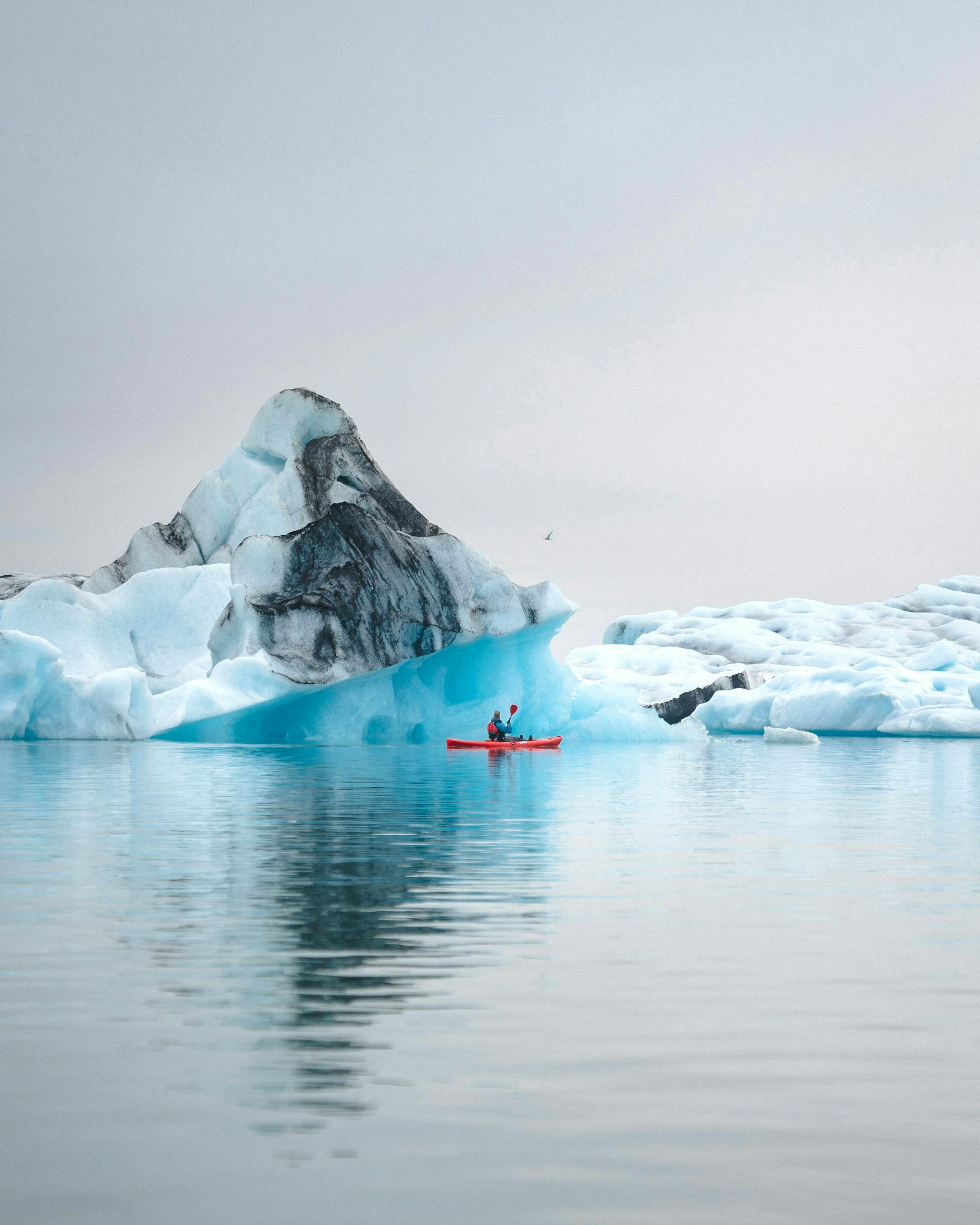 Towering icebergs rise from calm water beneath a cloudy Antarctic sky, with distant mountains on the horizon.