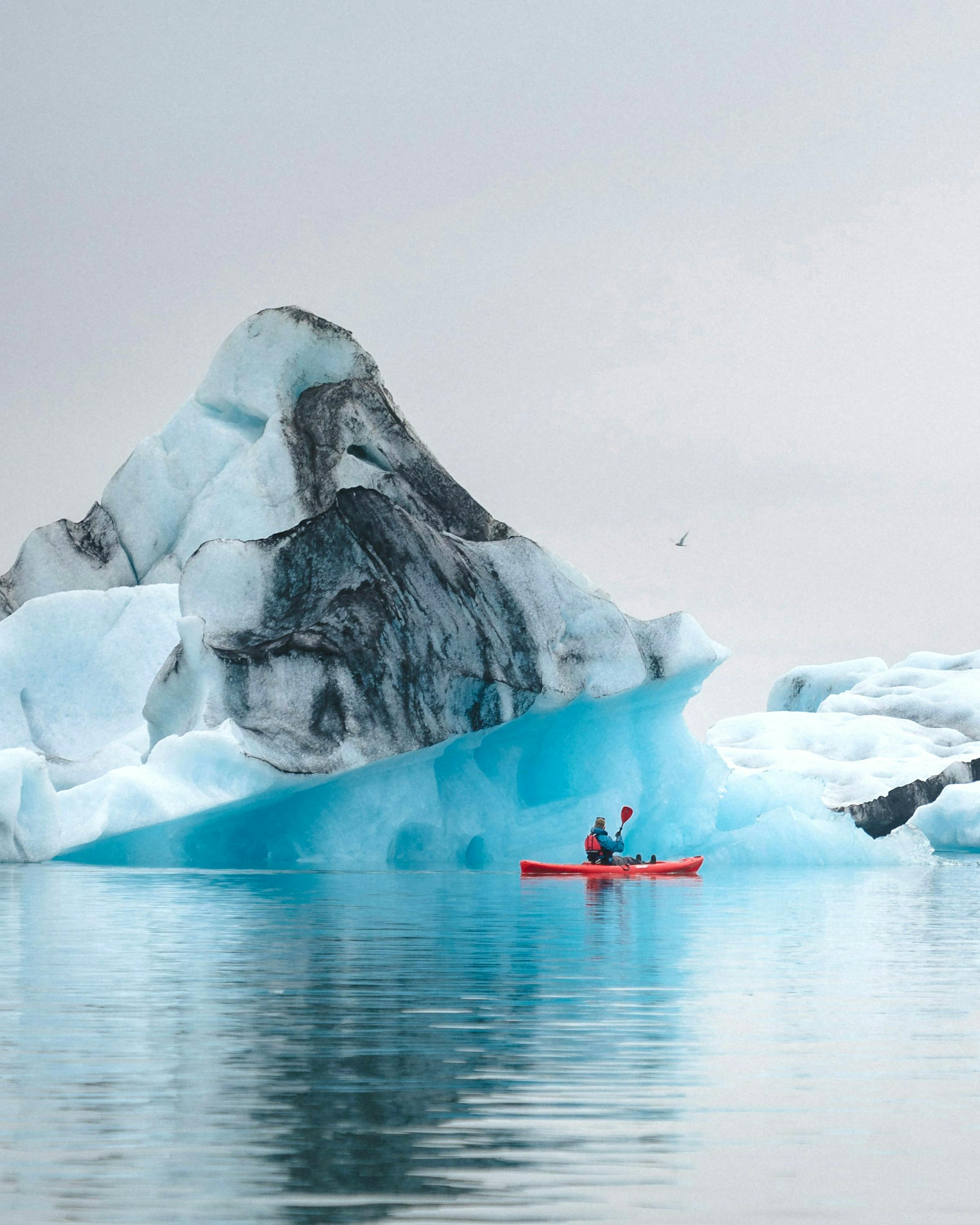 Towering icebergs rise from calm water beneath a cloudy Antarctic sky, with distant mountains on the horizon.
