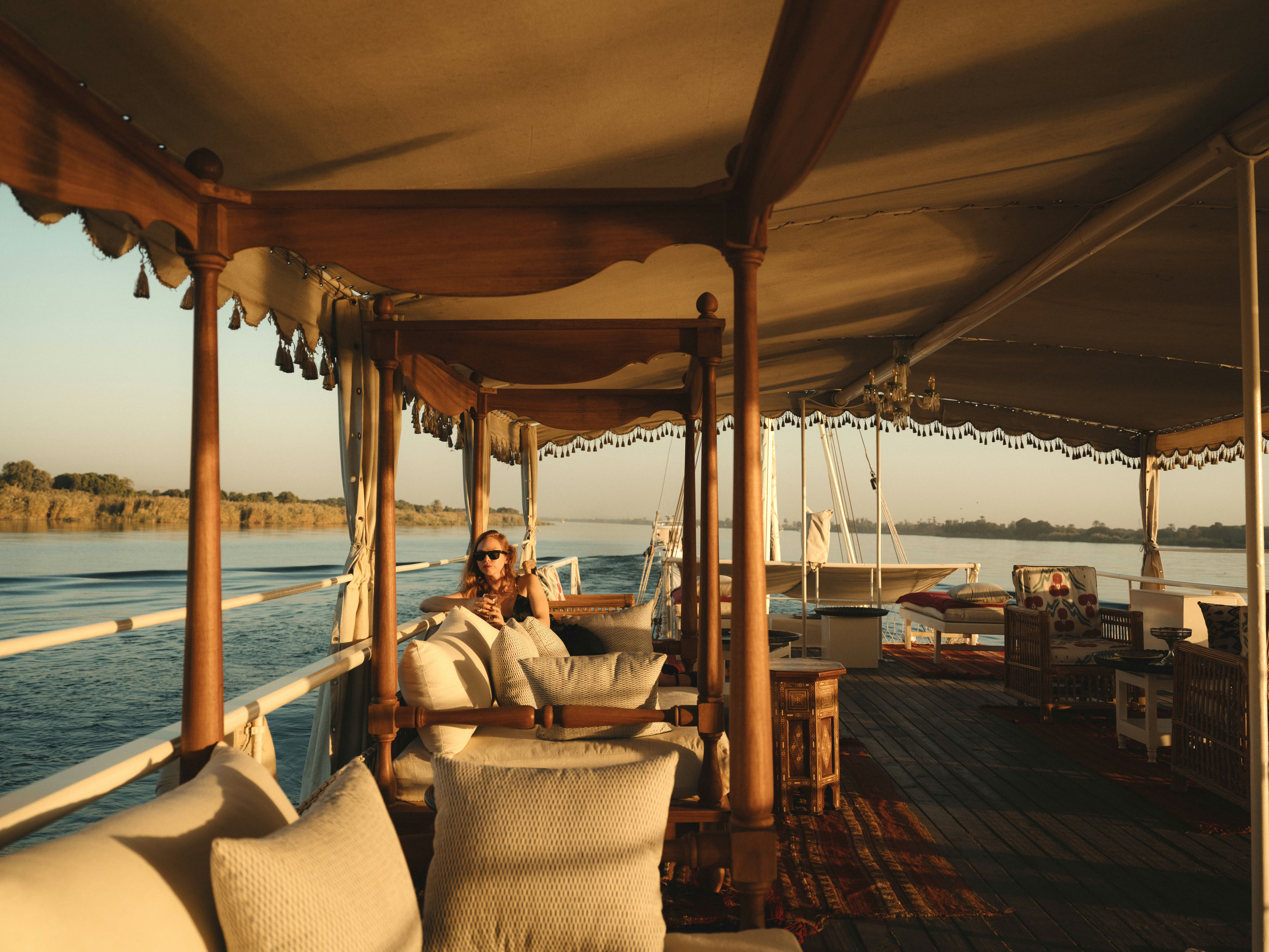 A person reclines on a cushioned chair on a shaded boat deck beside blue water and a sandy shoreline.