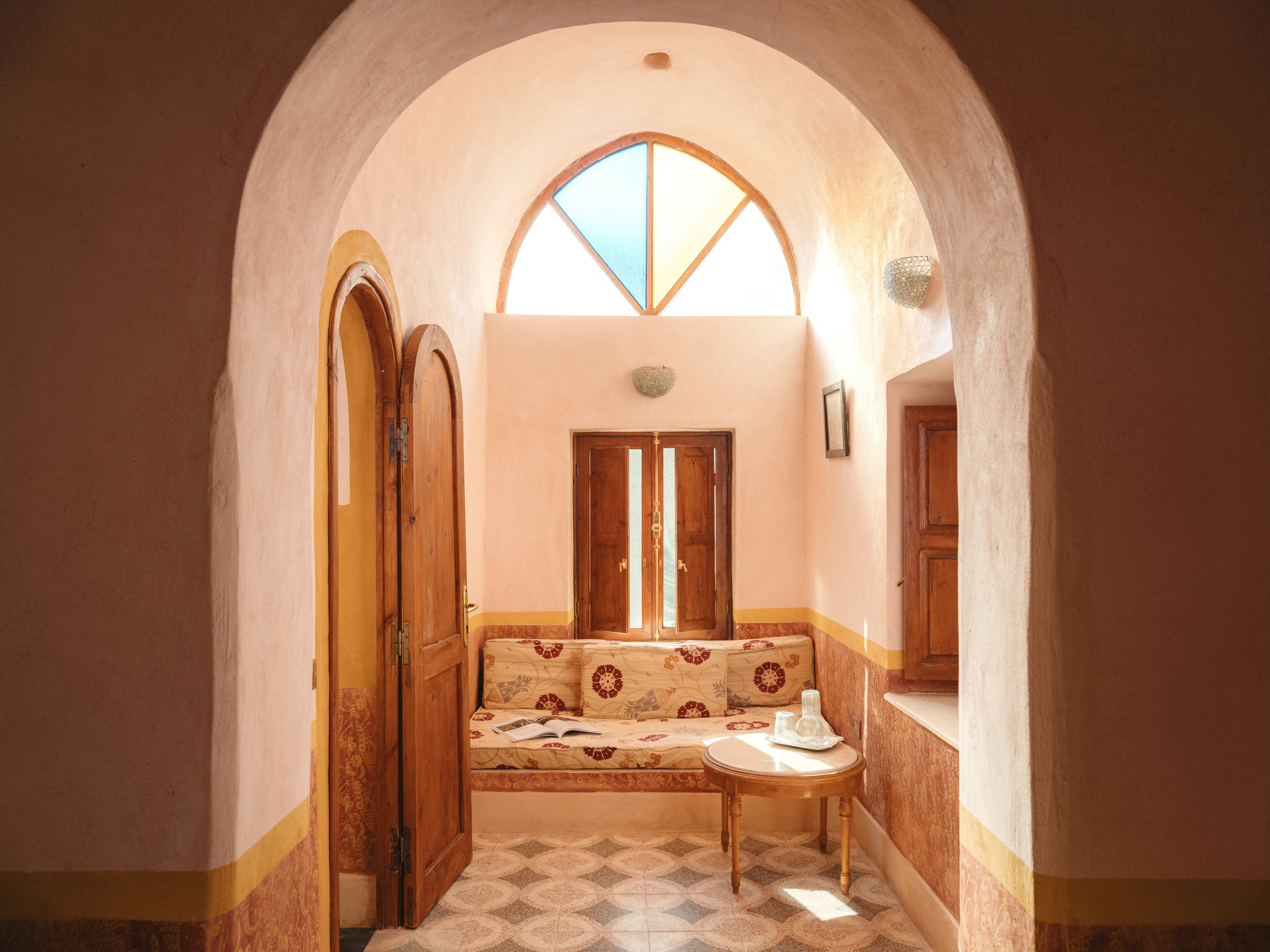 Sunlit arched doorway opens to a room with patterned tile floors and a single bed.