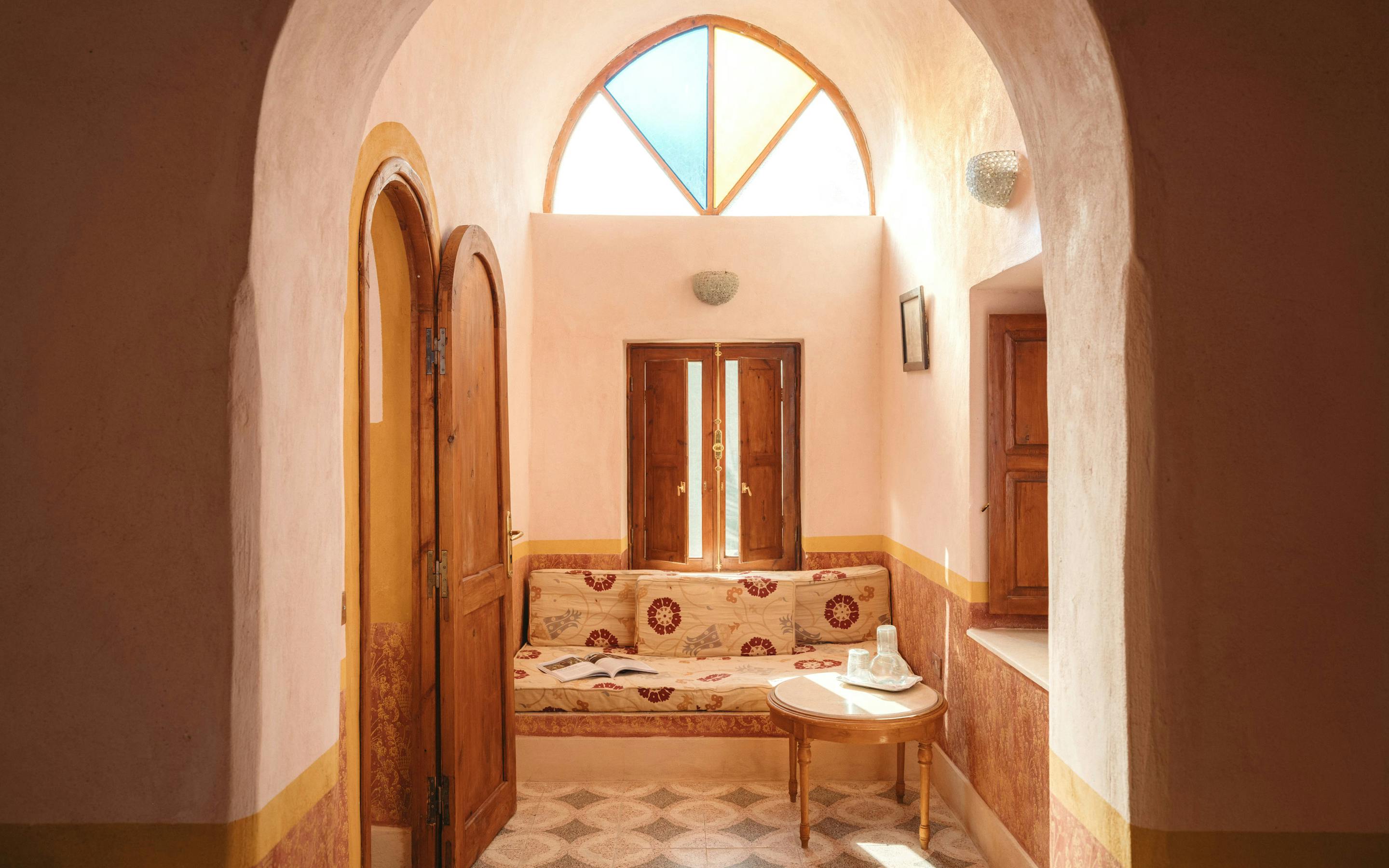 Sunlit arched doorway opens to a room with patterned tile floors and a single bed.