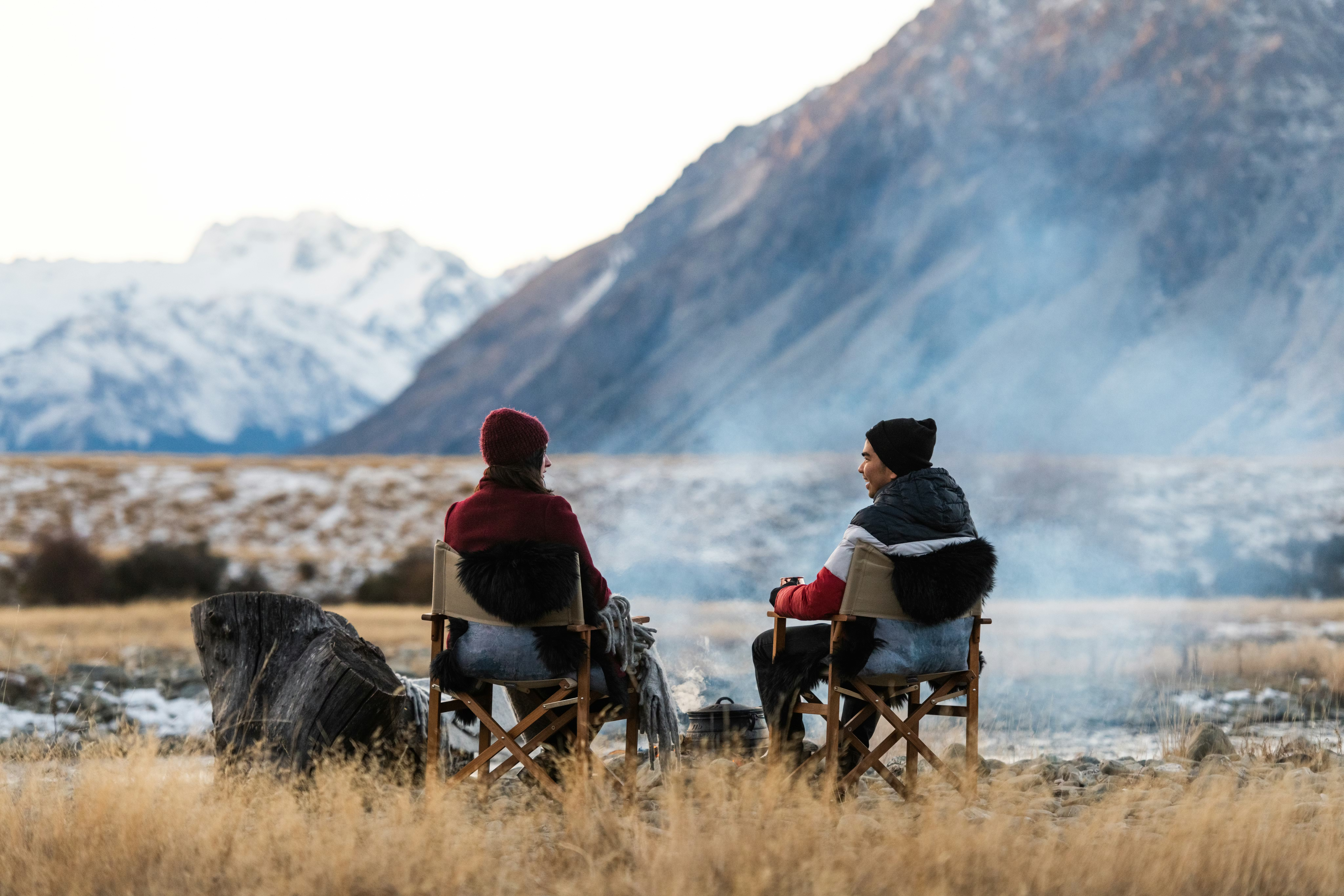 Two people sit by a small campfire on dry grass, holding warm drinks while snowy mountains glow in the distance.