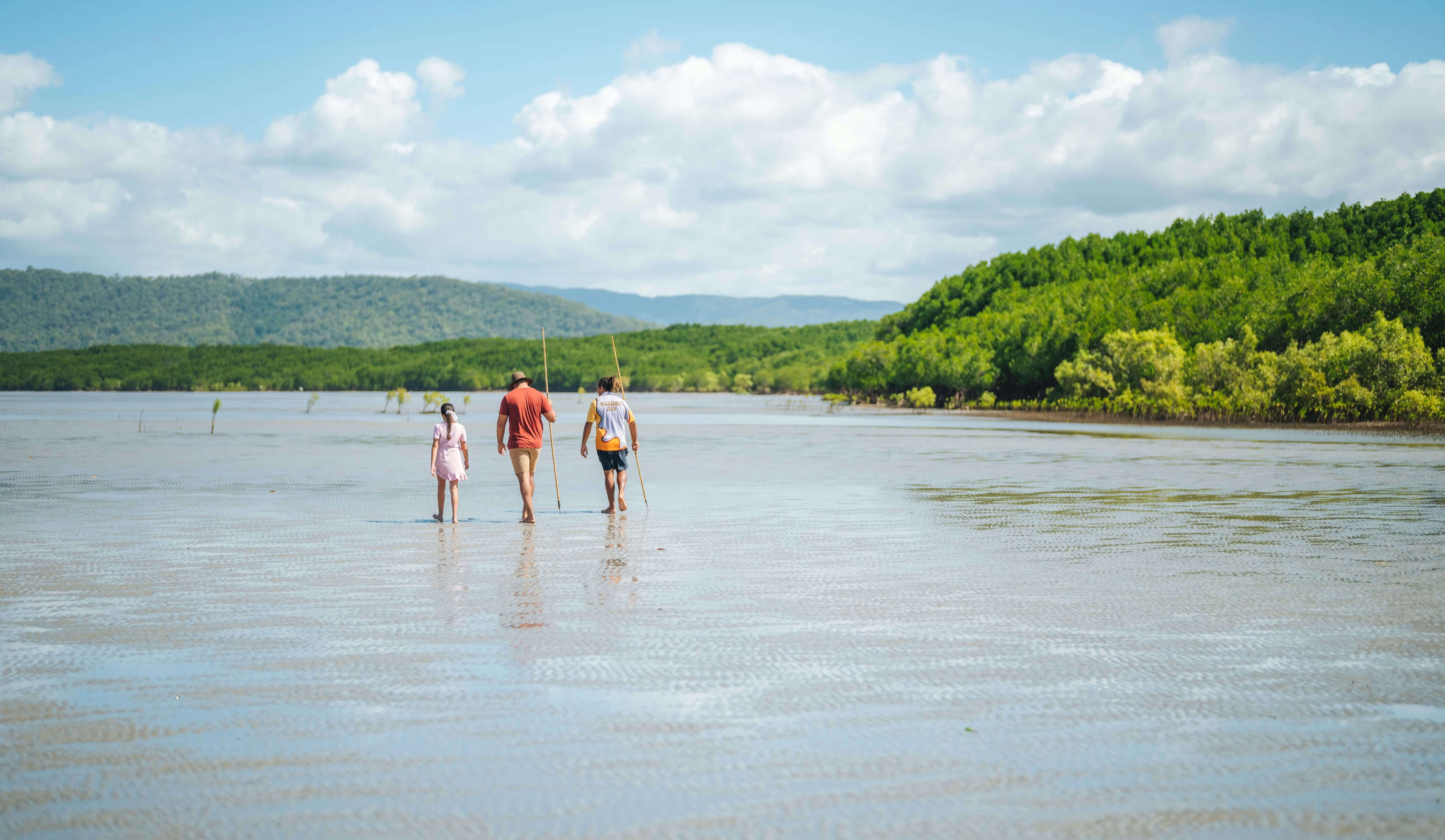 Three travelers wade through shallow water toward green mangroves, with low hills and clouds on the horizon.
