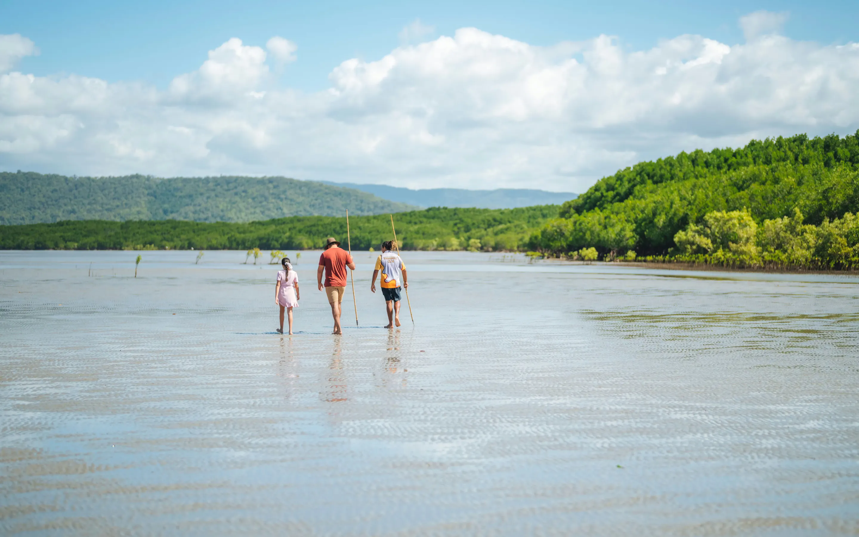 Three travelers wade through shallow water toward green mangroves, with low hills and clouds on the horizon.