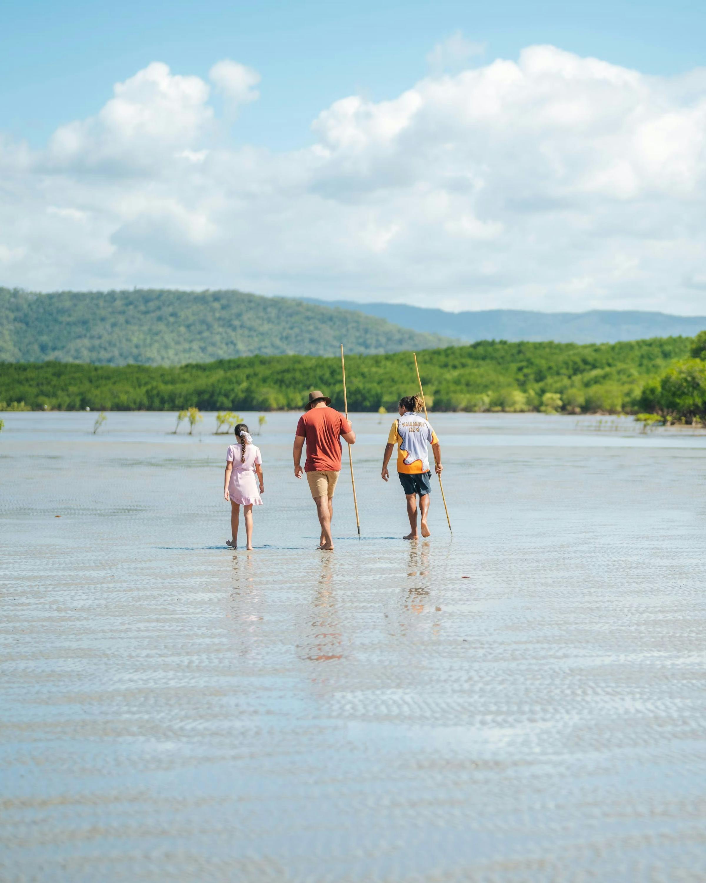 Three travelers wade through shallow water toward green mangroves, with low hills and clouds on the horizon.