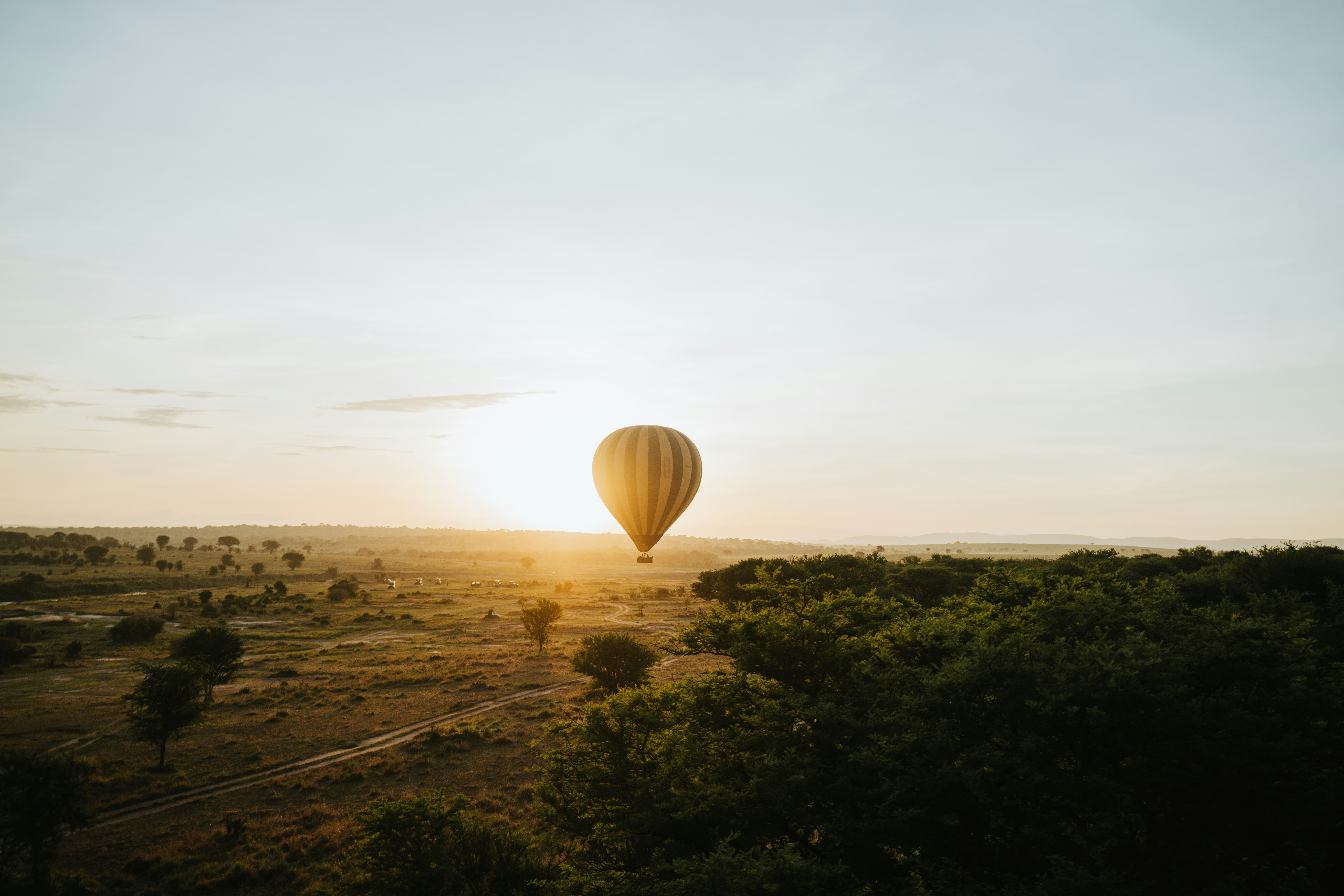 A hot air balloon floats above sunlit savanna at dawn, casting long shadows across trees and open plains.