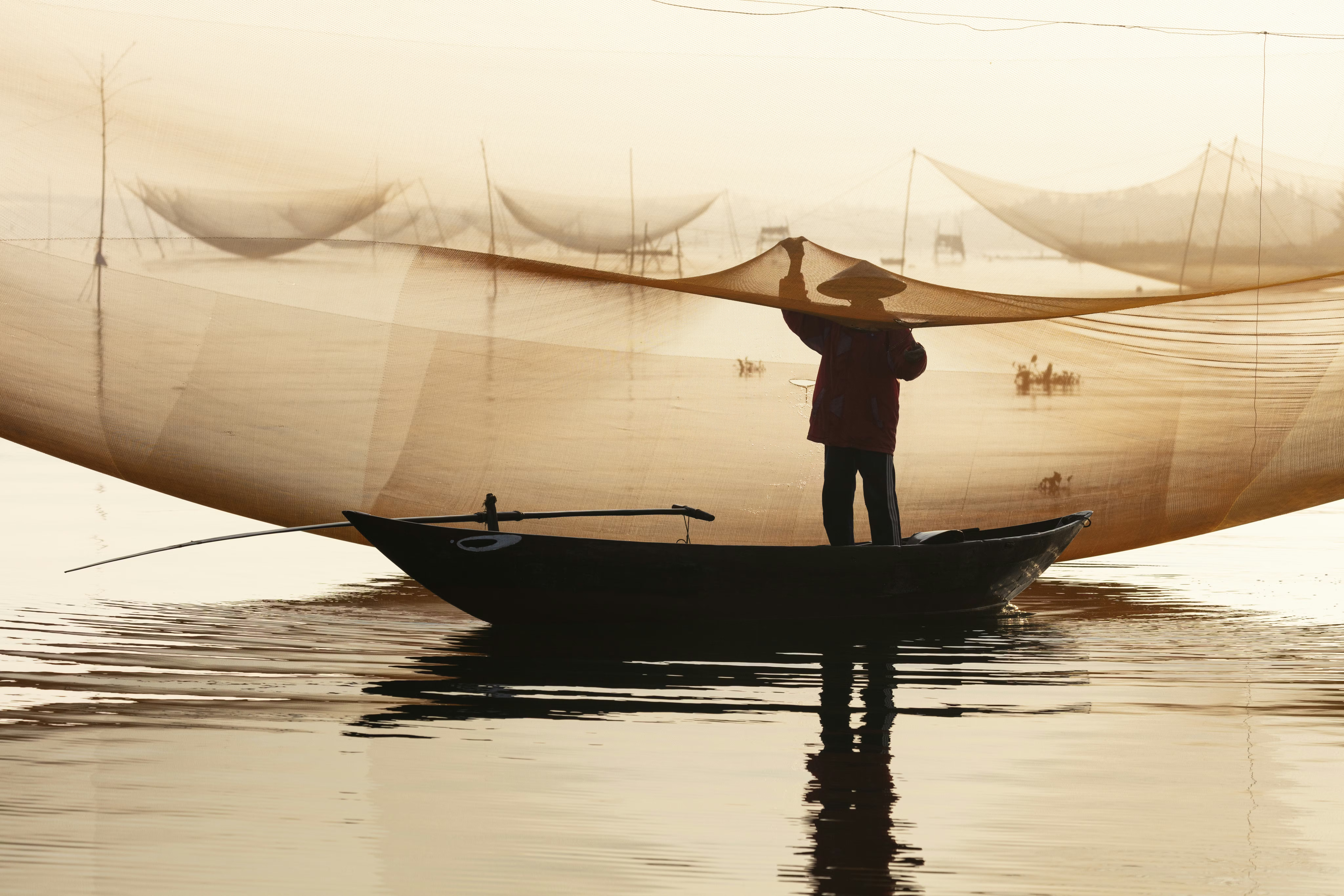 Person on a boat holds fishing nets over calm water at sunrise.